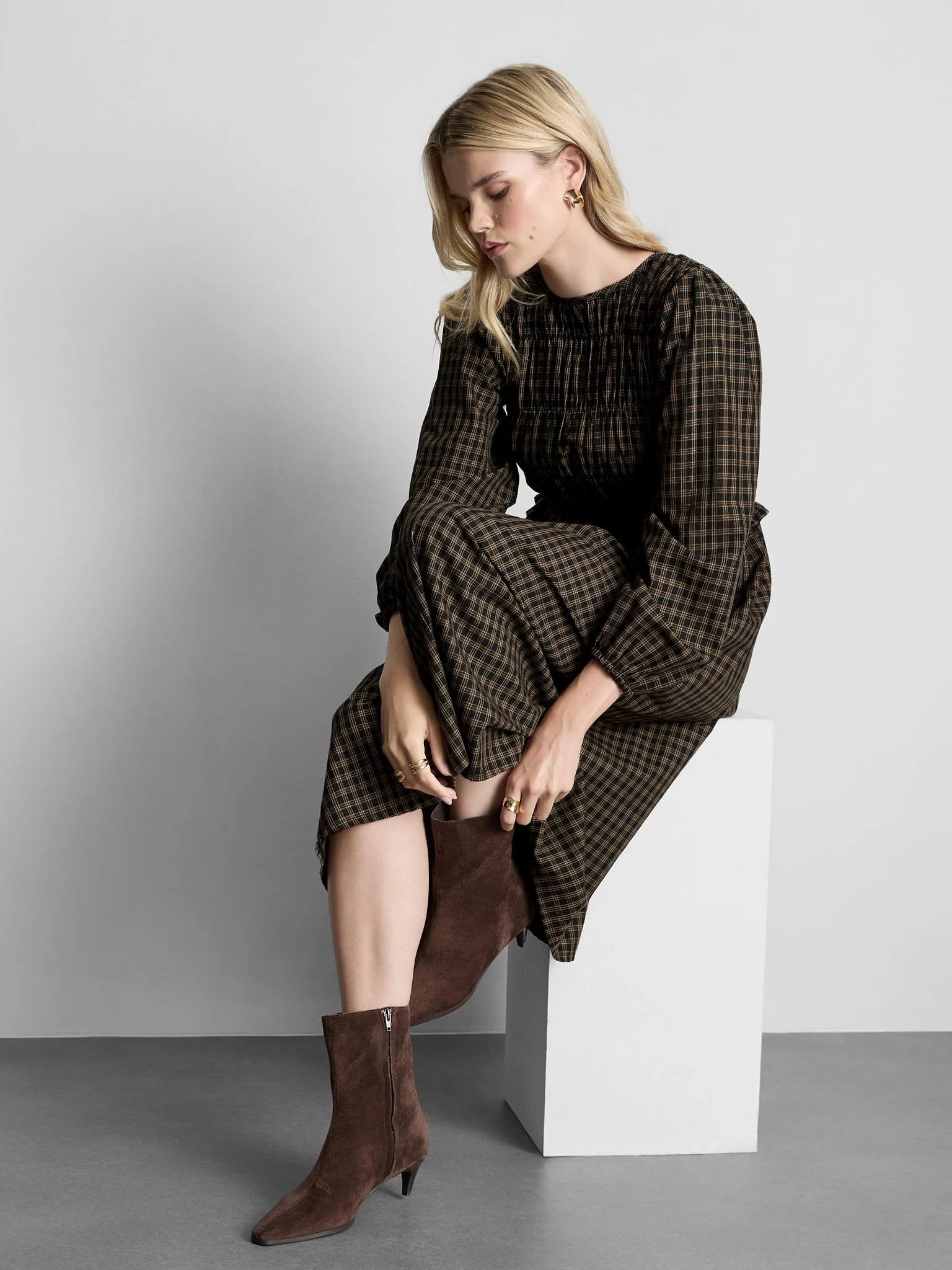 A woman with blonde hair wearing a brown plaid dress and brown knee-high boots, sitting on a white block against a plain background.