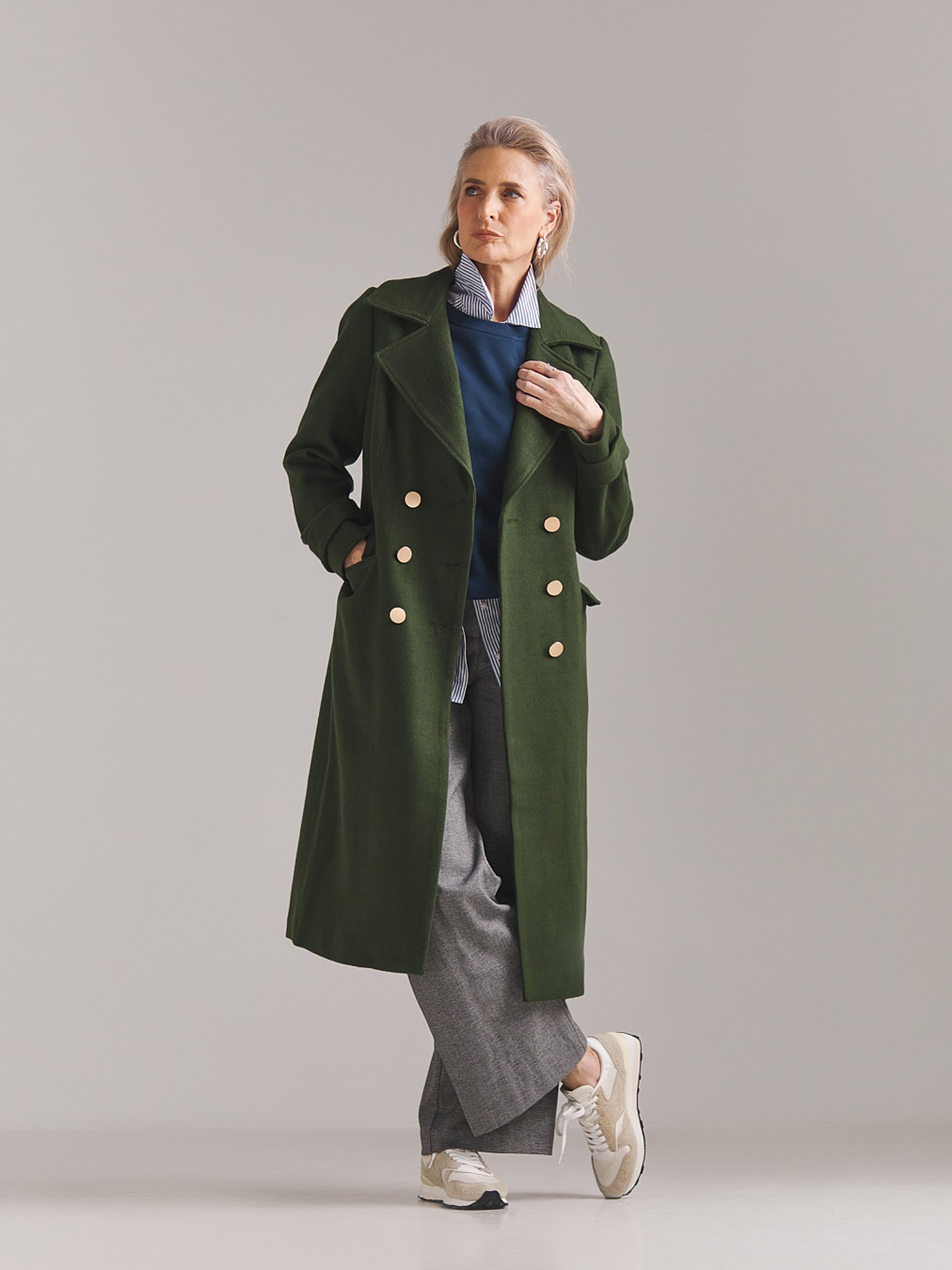 A woman wearing a long green double-breasted coat over a blue shirt, gray pants, and beige sneakers, standing against a plain light gray background.