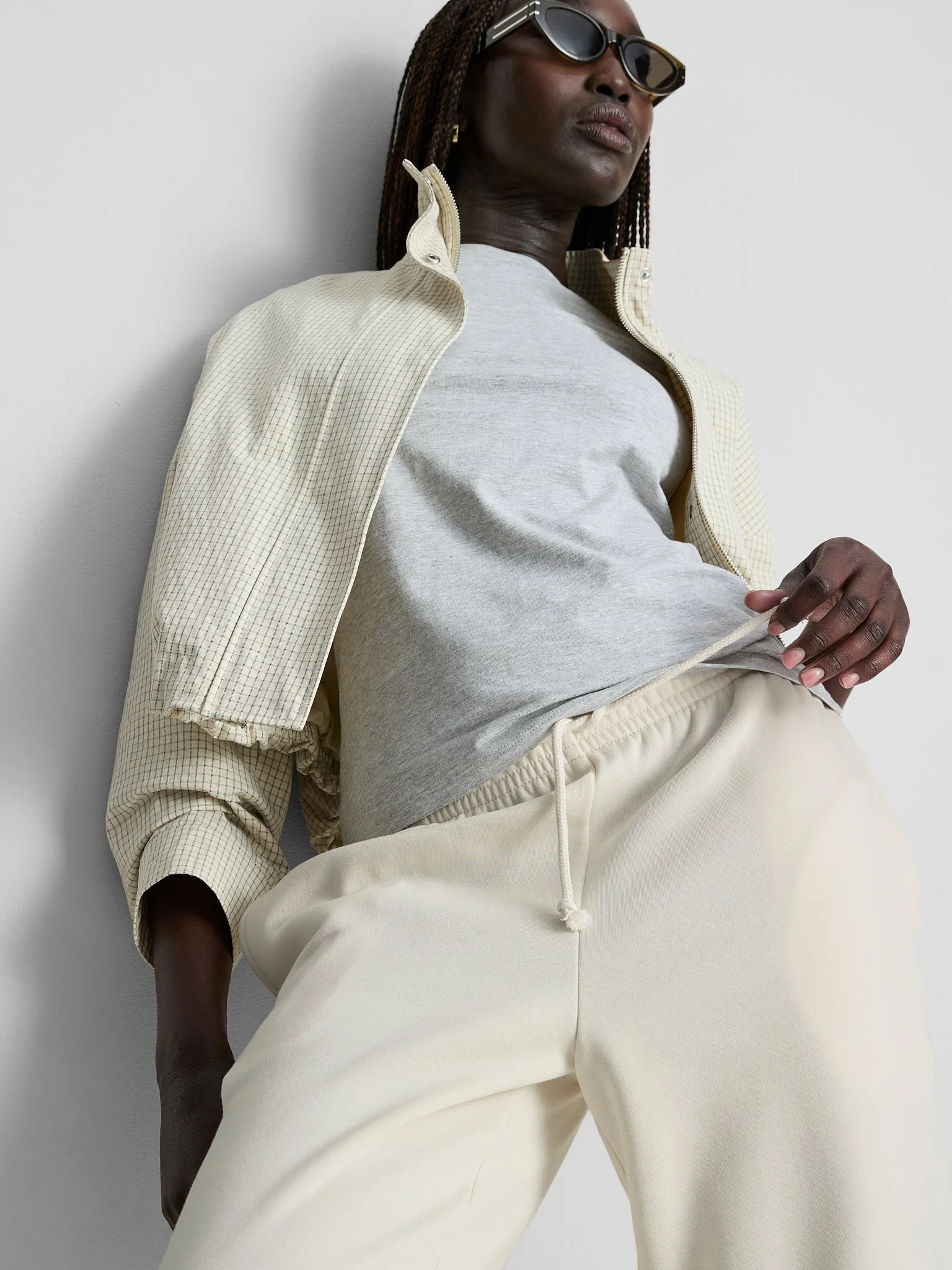 A woman with dark skin, wearing black sunglasses, a light grey t-shirt, a light beige zip-up jacket with a checkered pattern, and cream-colored pants posing against a plain white wall.