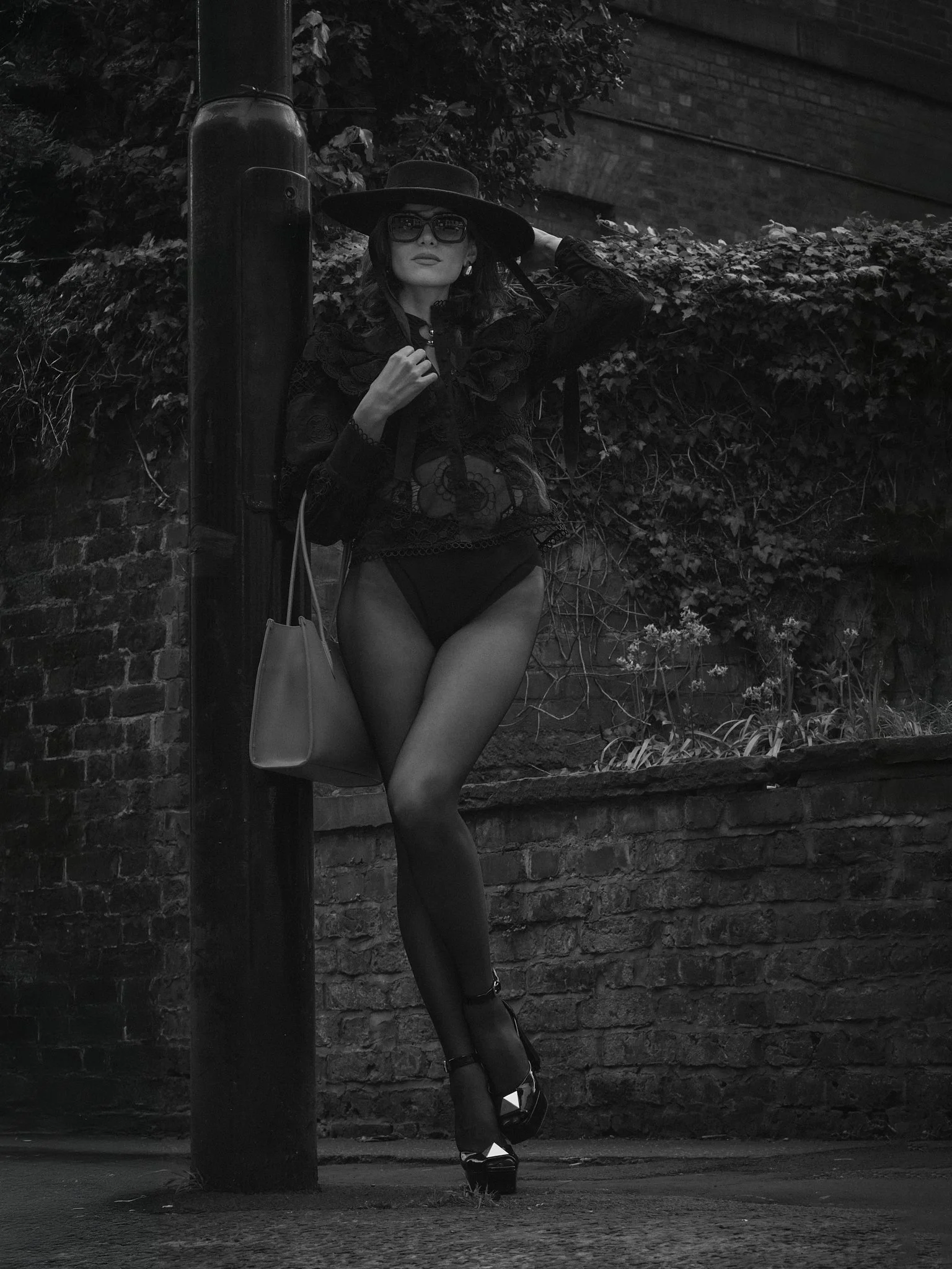 A woman in a black lace top
and shorts, wearing a large hat and sunglasses, leaning against a brick wall behind a pole