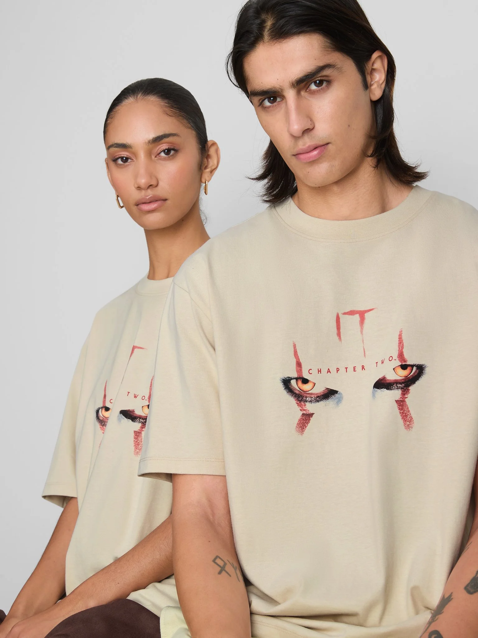 Three young adults with serious expressions, wearing cream-colored T-shirts with a graphic of menacing eyes and the text 'Chapter Two,' posing against a plain light gray background.