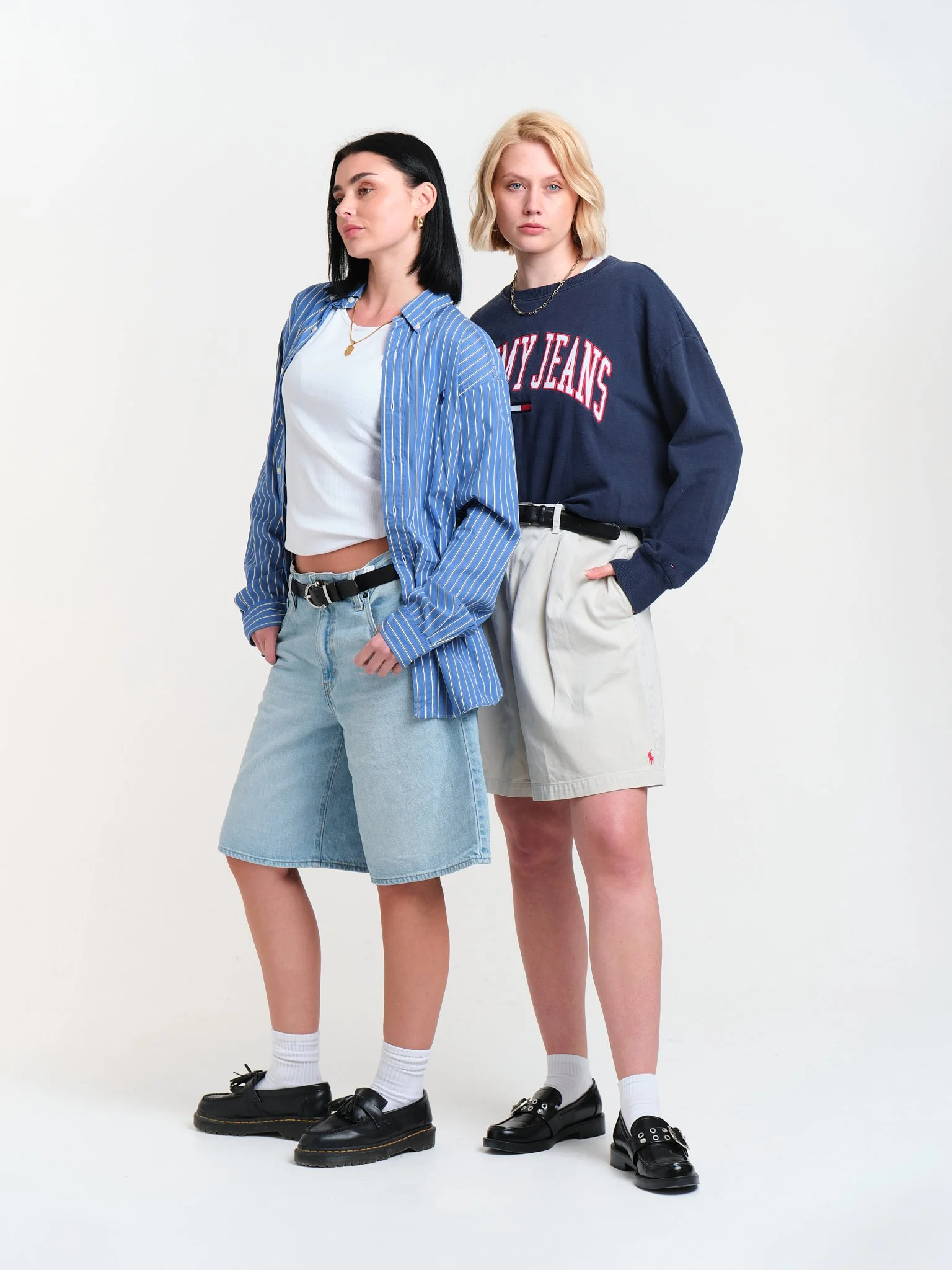 Two young women standing against a plain white background, wearing casual clothing. The woman on the left has black hair, a blue and white striped shirt, a white crop top, denim skirt, black shoes, and white socks. The woman on the right has blonde h