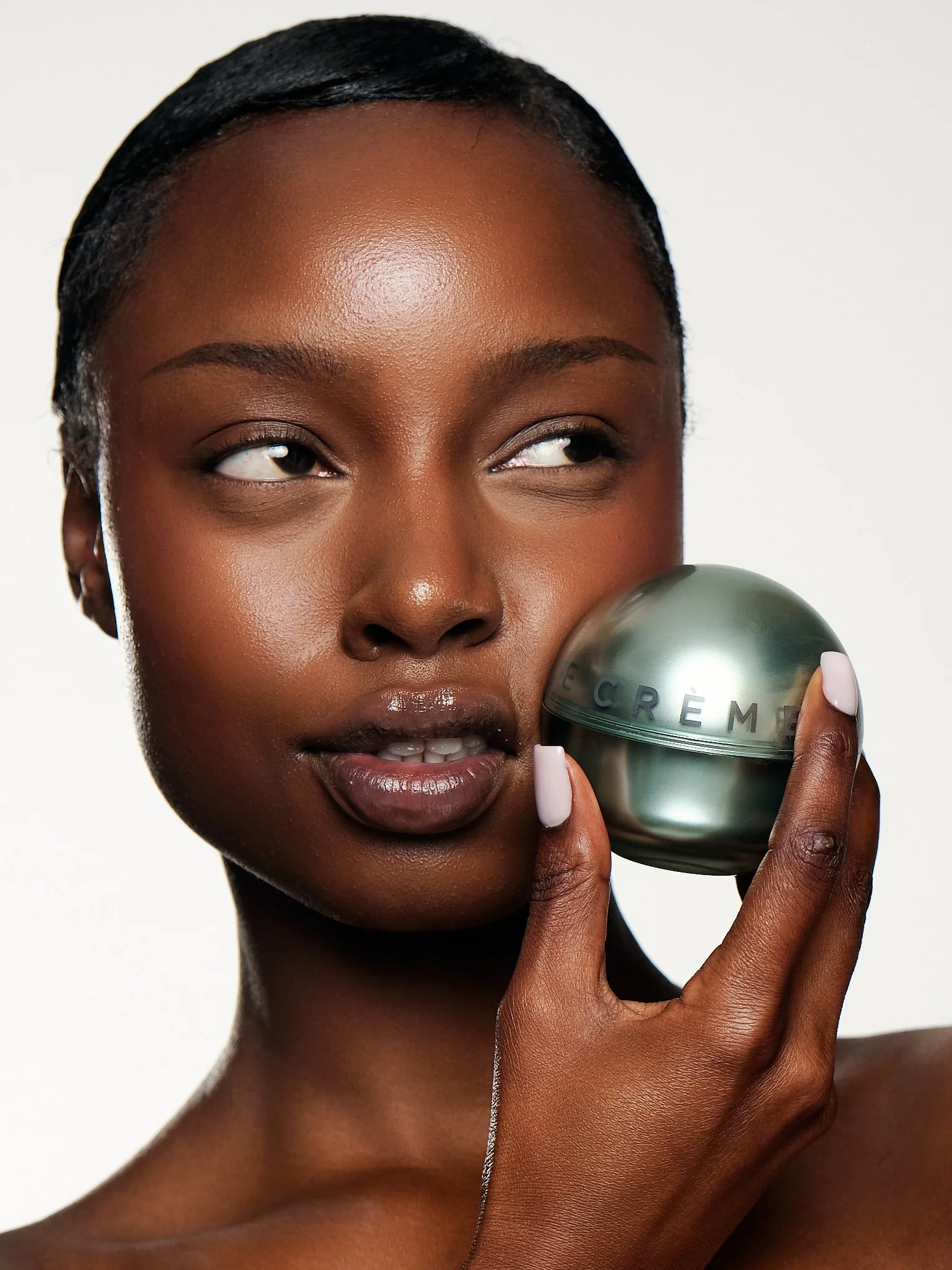 A young woman with dark skin and short black hair holds a metallic skincare product near her lips, with a neutral white background.
