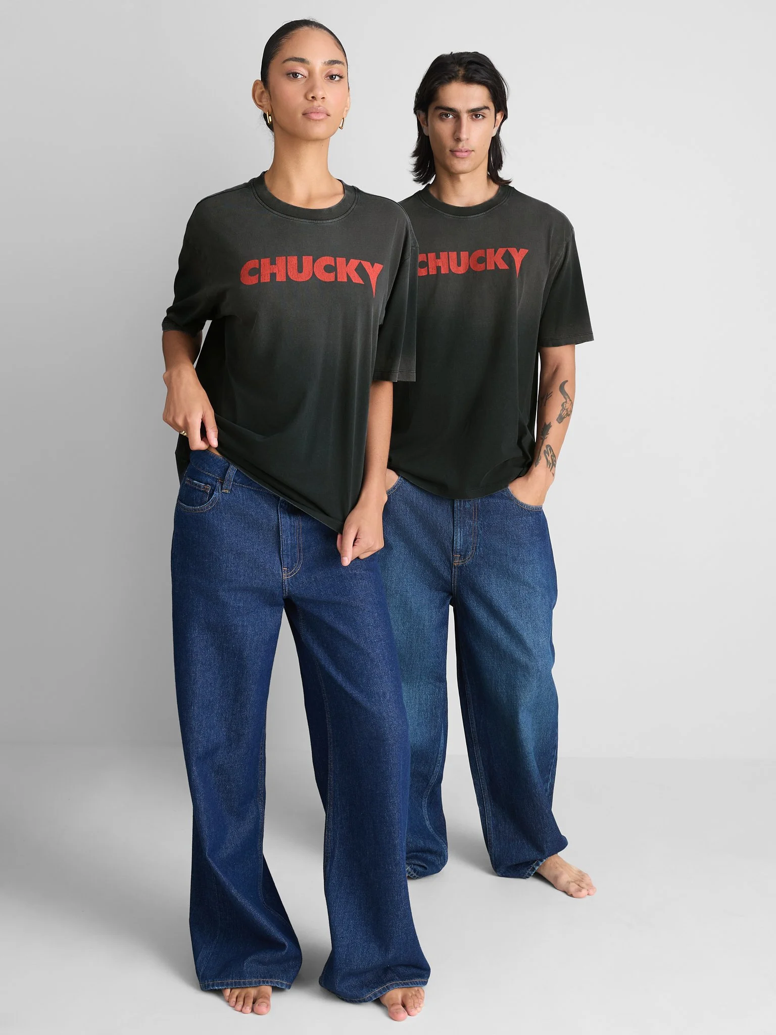Two individuals standing side by side, both wearing black T-shirts with the word 'CHUCKY' in red across the front, and blue jeans. The person on the left has their hair pulled back and is looking directly at the camera, while the person on the right 