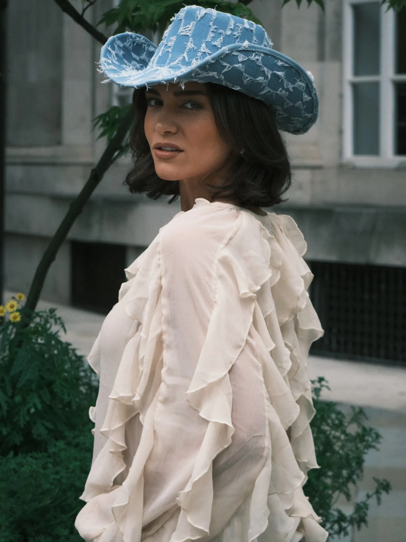 A woman with dark hair wearing a large, distressed, light blue cowboy hat standing outdoors near green plants and a building.