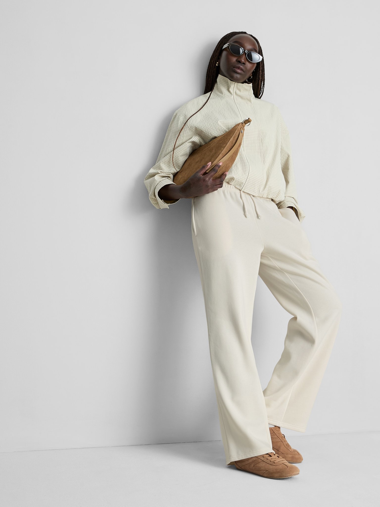 A woman with dark skin, wearing sunglasses, a beige cropped jacket, cream-colored sweatpants, and beige sneakers, standing against a plain white wall, holding a tan crossbody bag.