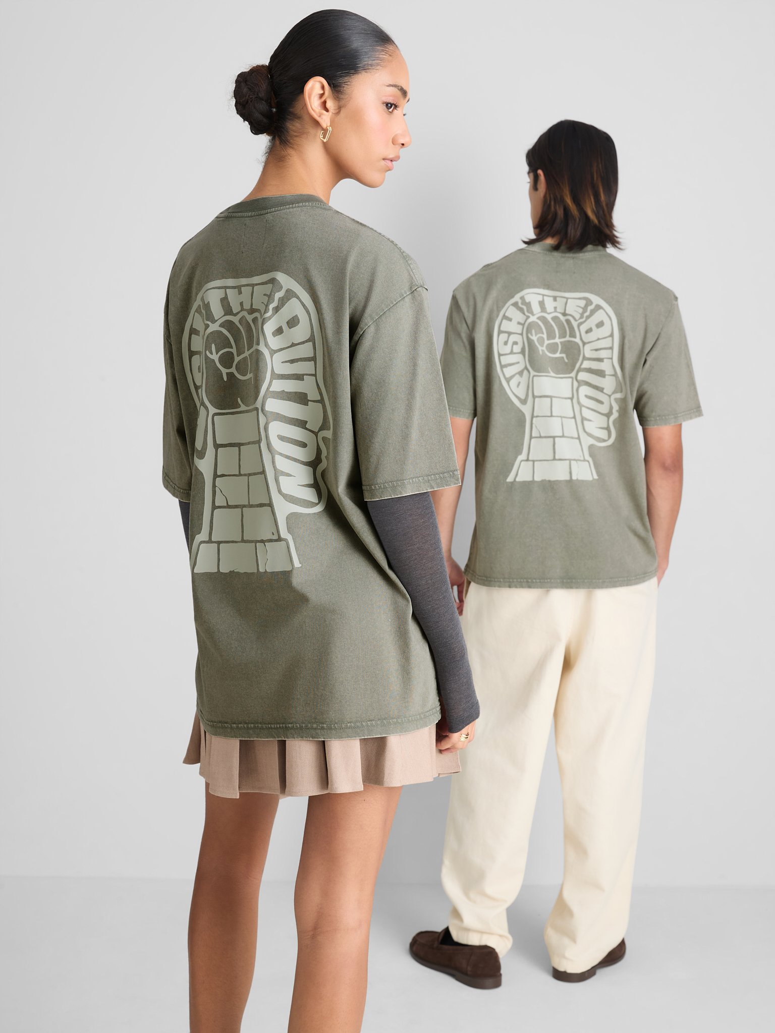 Two women standing with their backs to the camera, wearing loose-fitting gray T-shirts with a large graphic of a raised fist and the words "The Boycott" on the back. One woman has long dark hair in a bun, and the other has shoulder-length hair.