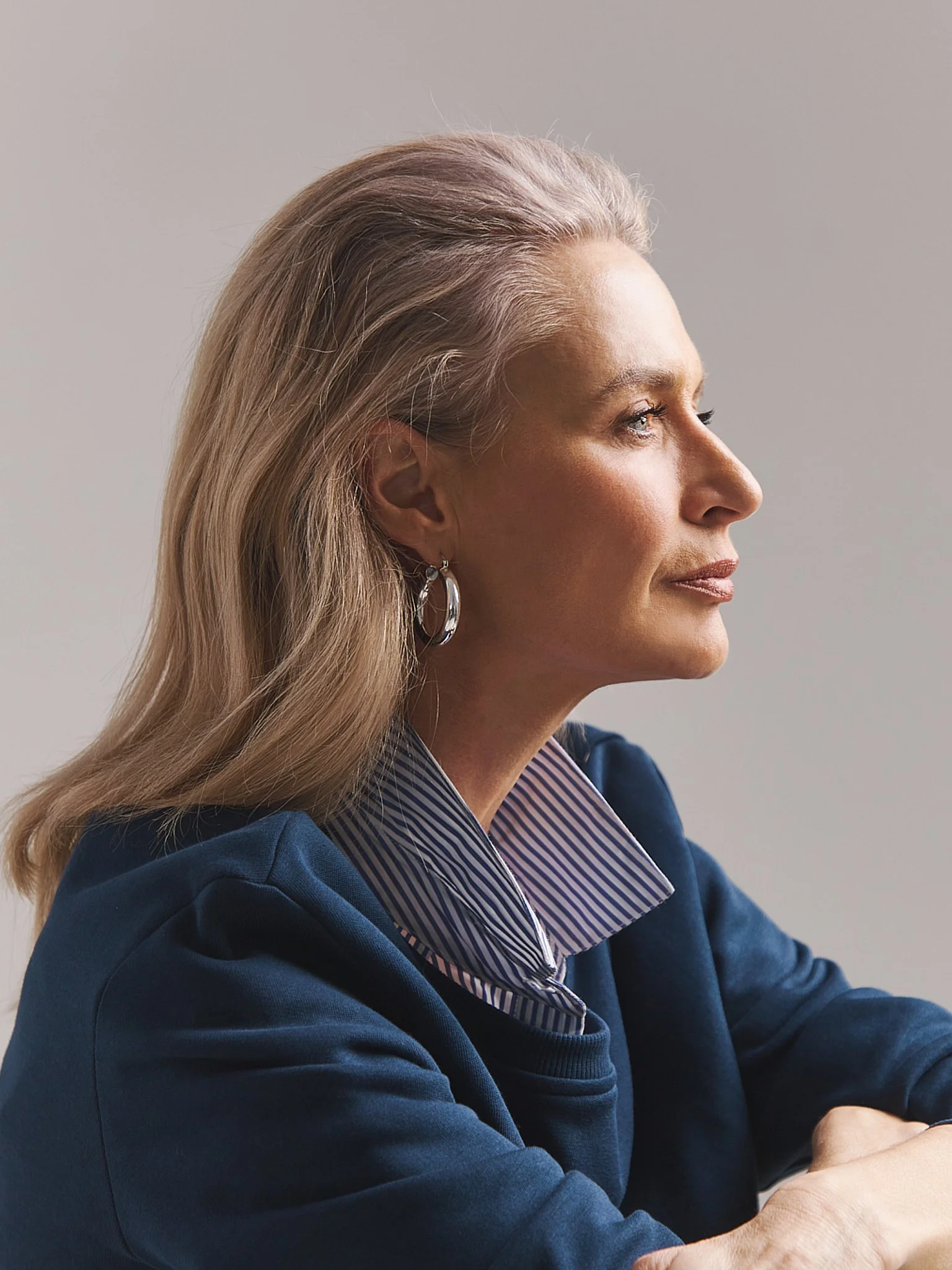 Profile of a woman with blonde hair, wearing a blue blazer and a striped shirt, looking to her right, with a neutral background.