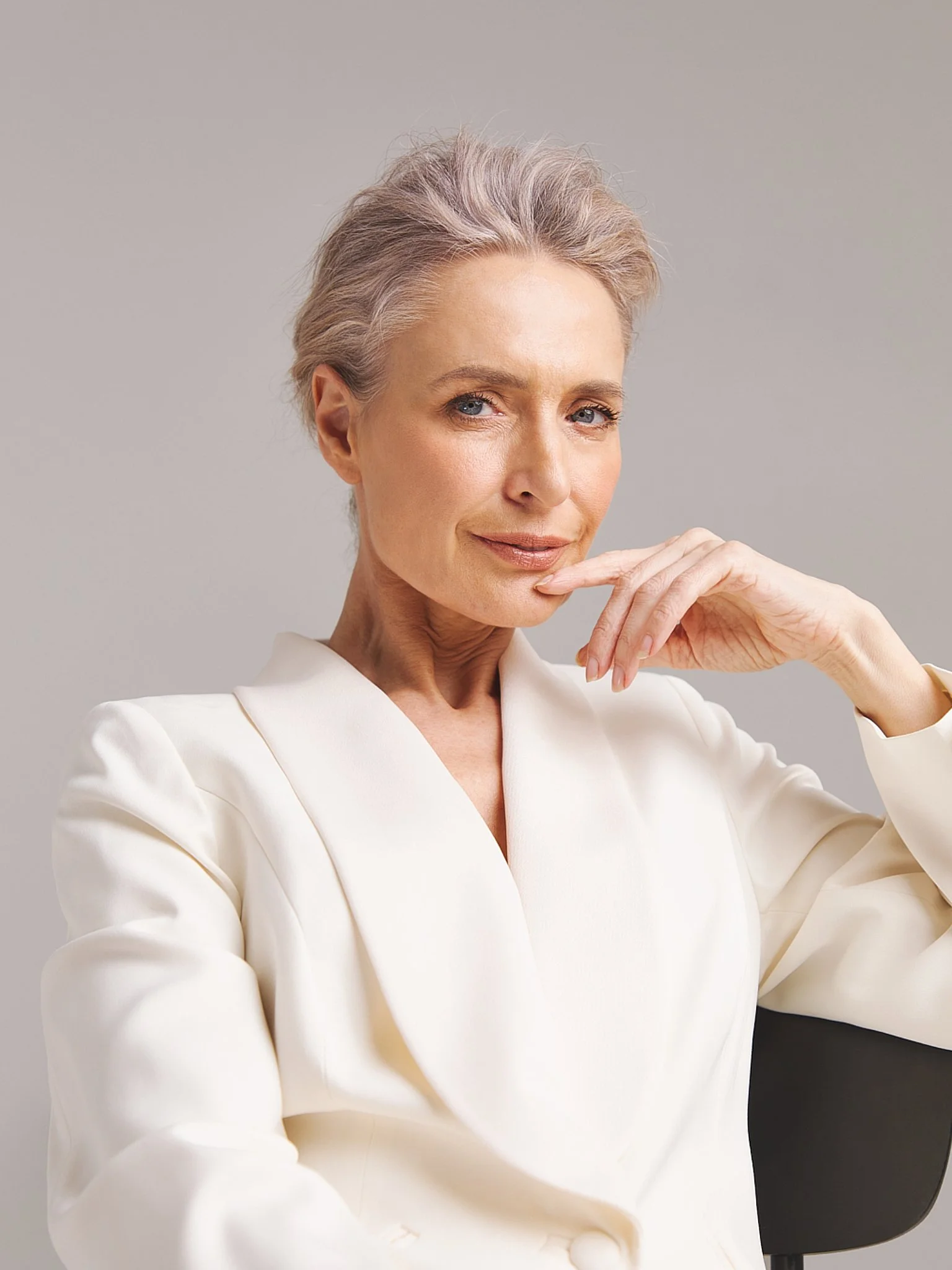 An elegant older woman with short, gray hair, wearing a white blazer, sitting on a black chair, resting her chin on her hand, looking confidently at the camera against a plain gray background.