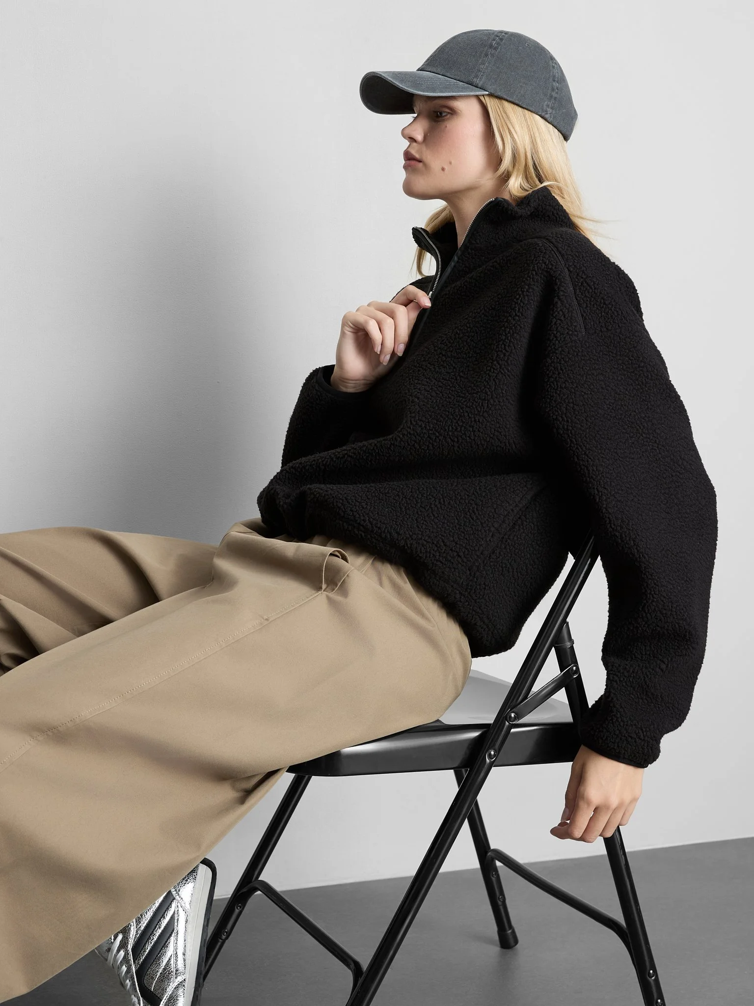 A young woman wearing a dark cap, black sweater, and tan pants, seated on a black foldable chair with a neutral gray background.