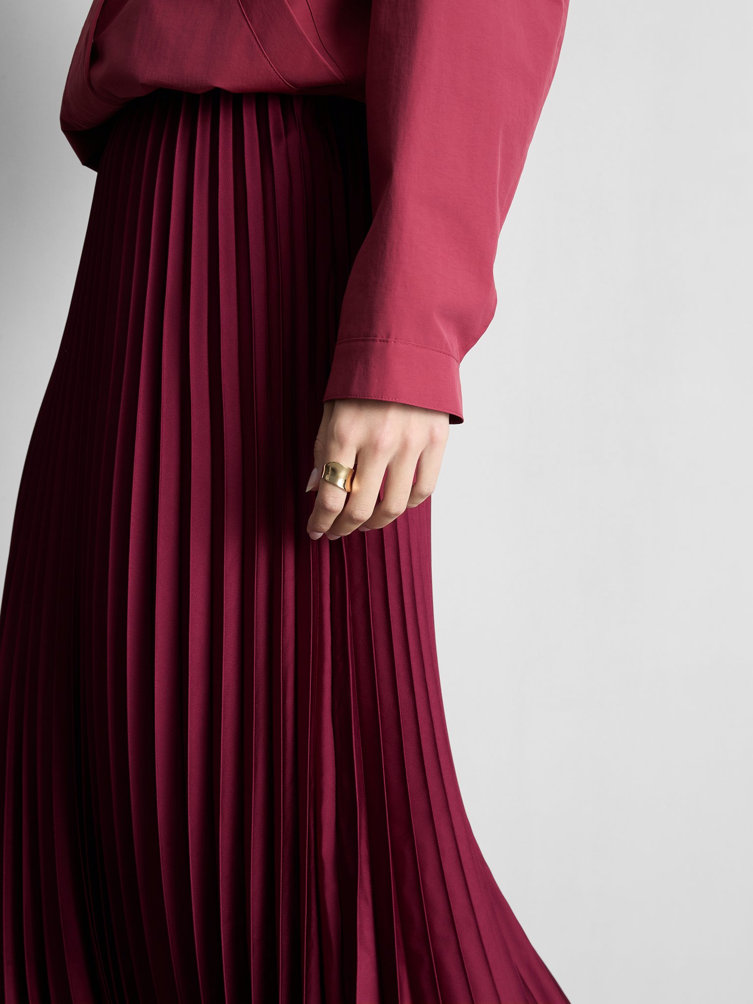 Close-up of a person wearing a long, pleated maroon skirt and a matching maroon shirt, with a gold ring on their finger, against a plain white background.