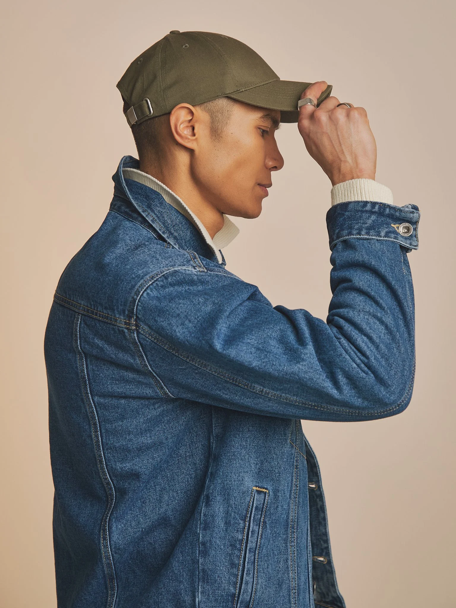 Profile of a young man wearing a green baseball cap, a denim jacket, and a white sweater underneath, with his eyes closed and head tilted slightly downward, holding the brim of his cap with one hand.