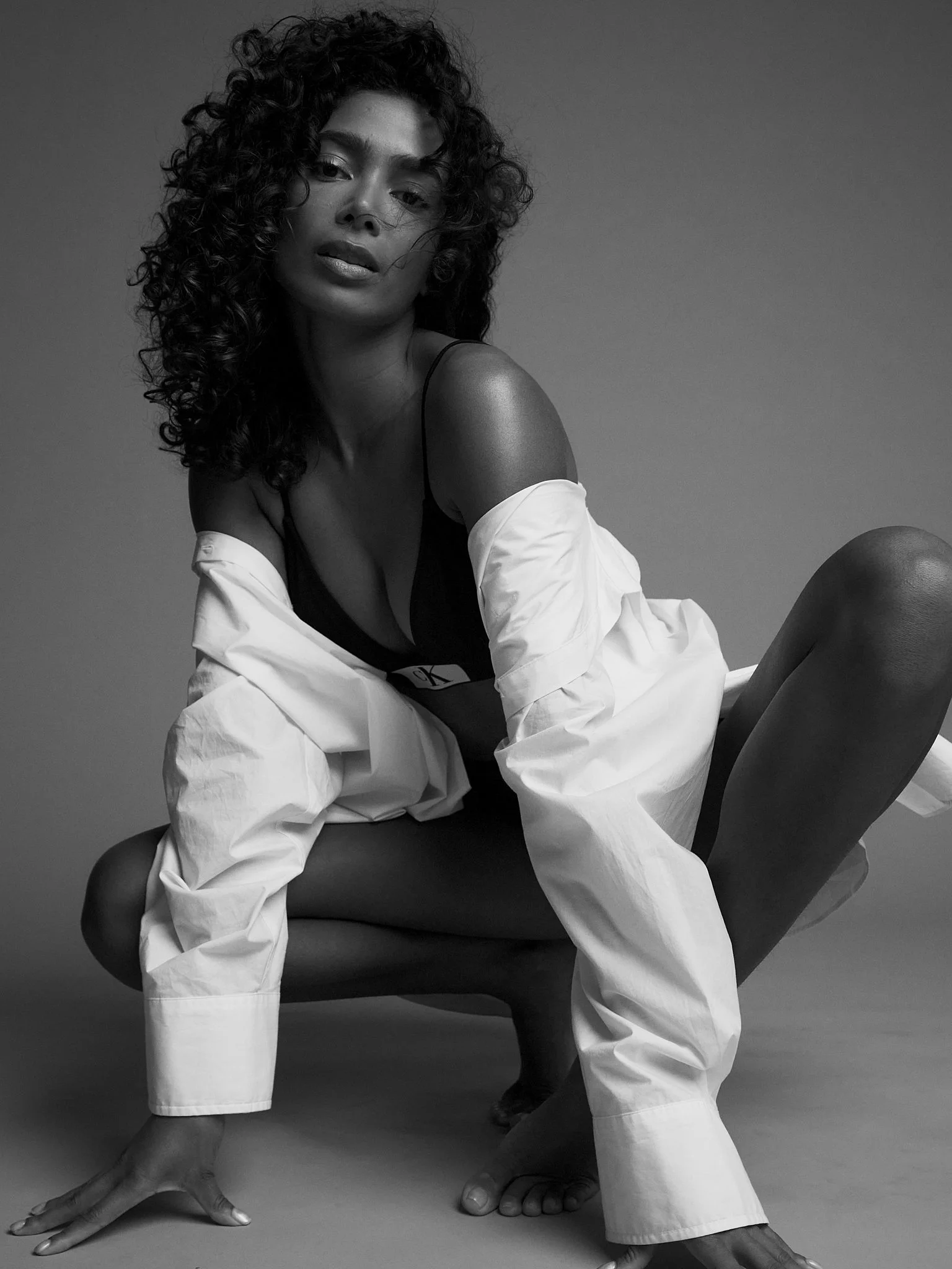 Black and white photo of a woman with curly hair, wearing a black top and white pants, sitting on the floor with her hands on the ground, looking at the camera.