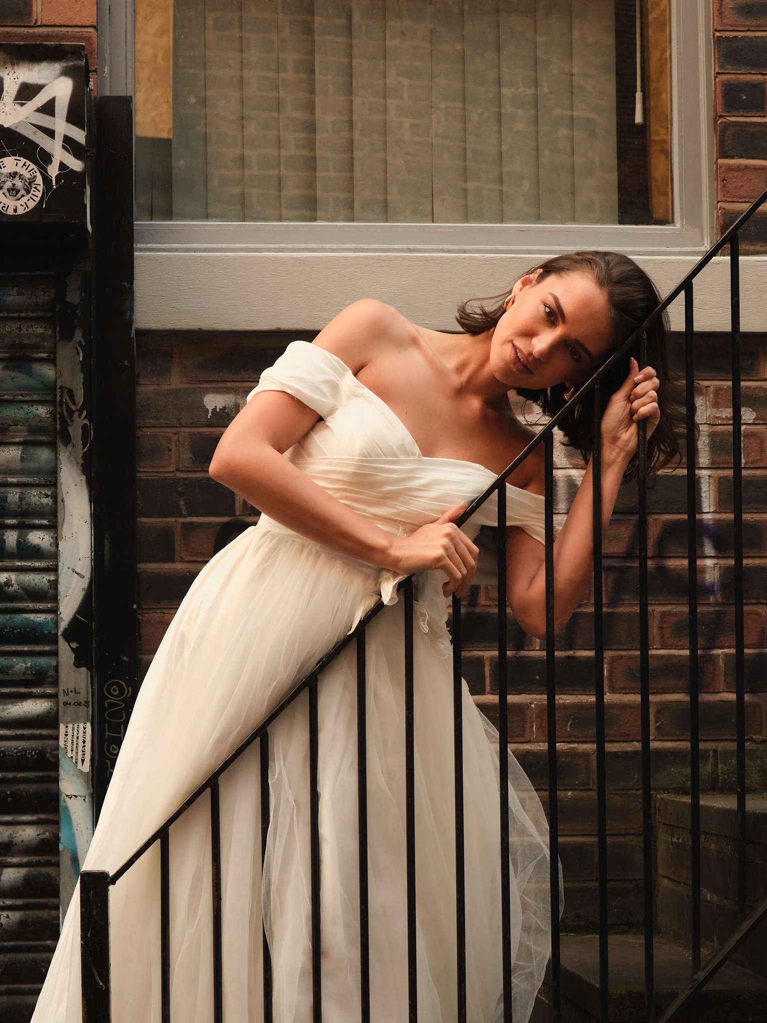 A woman wearing an off-shoulder white dress is leaning on a black metal railing on a stairway outside a brick building, smiling and looking at the camera.