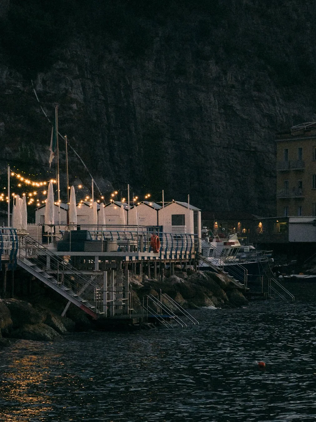 Nighttime scene of a waterfront with small buildings, string lights, boats, and stairs leading into the water, with a large rocky cliff in the background.