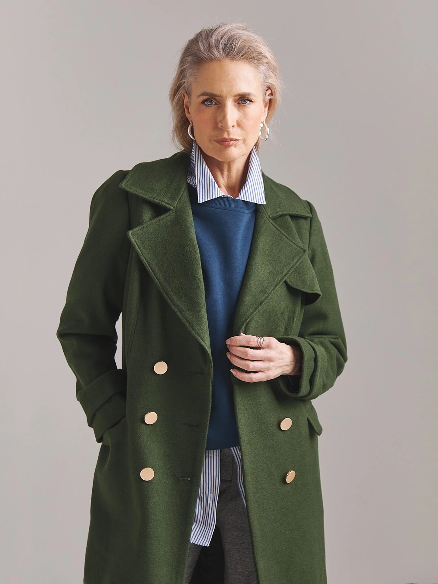 A woman with short blonde hair and hoop earrings, wearing a green coat over a blue sweater, striped shirt, and gray pants, standing against a plain gray background.
