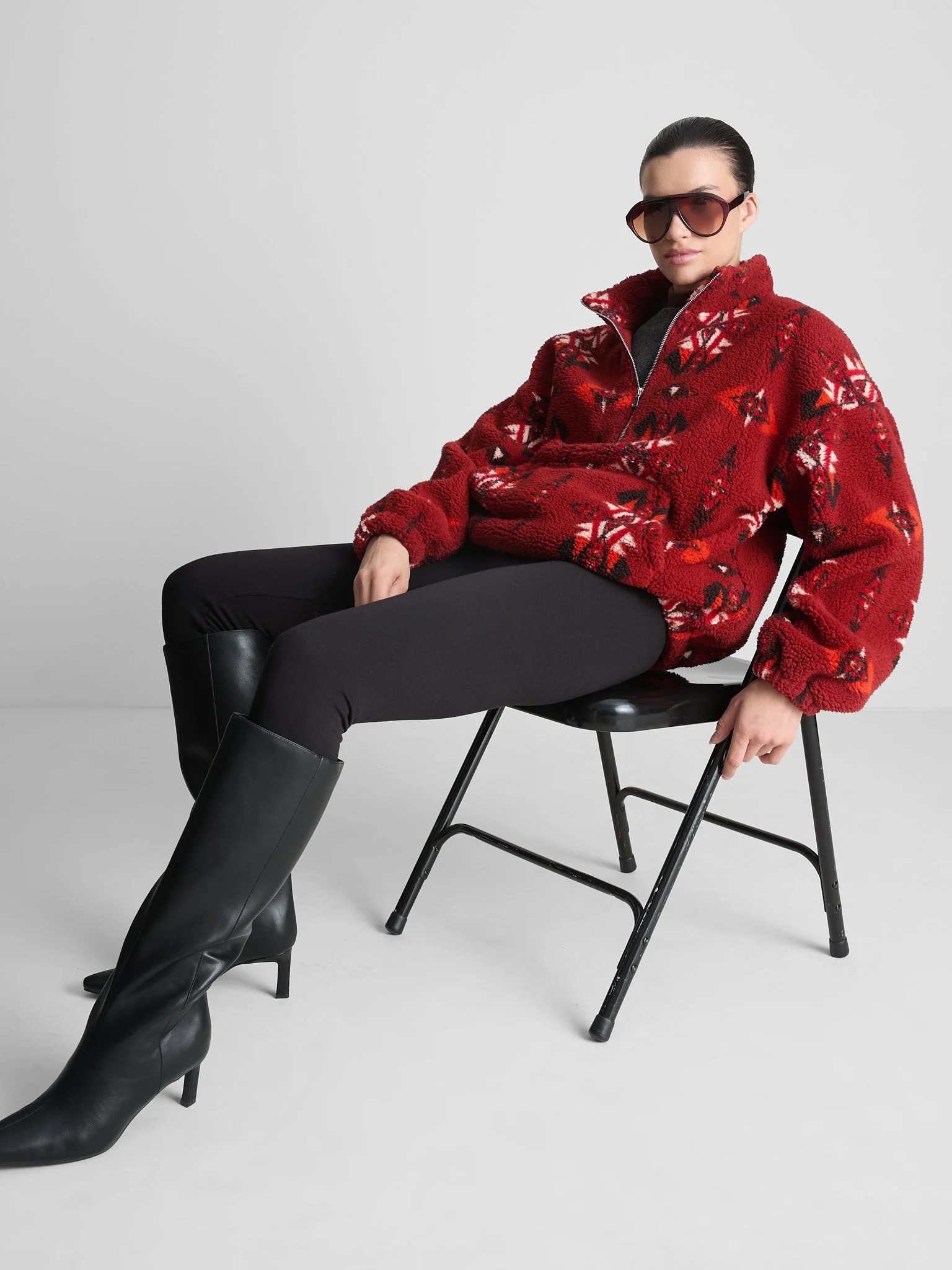 A woman with dark hair, wearing large sunglasses, a red patterned fleece jacket, black pants, and black boots, sitting on a black chair against a plain light gray background.