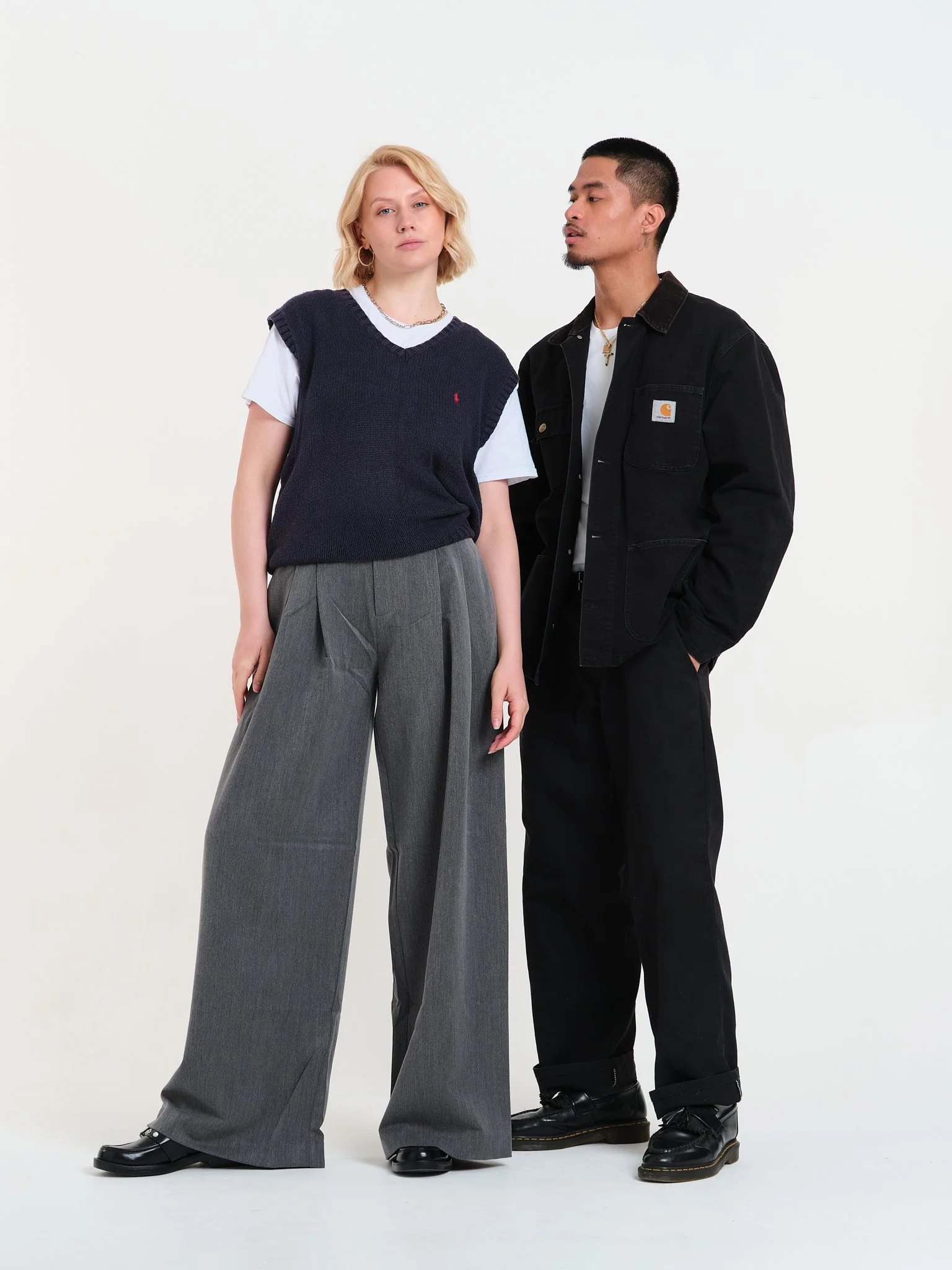 Two young adults standing against a plain white background, wearing casual streetwear. The woman has blonde hair, a navy vest over a white T-shirt, and wide-leg gray trousers. The man has dark hair, a black Carhartt jacket, dark pants, and boots, wea