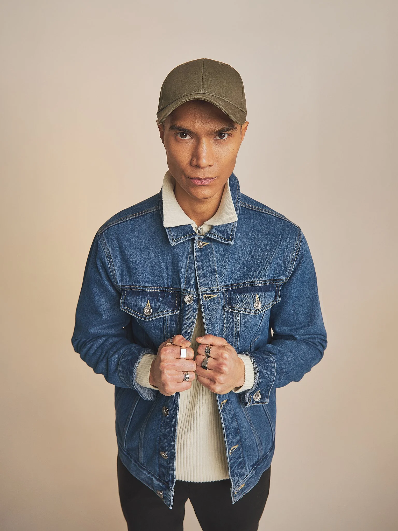 A person wearing a green cap, denim jacket, and rings, posing against a plain beige background.