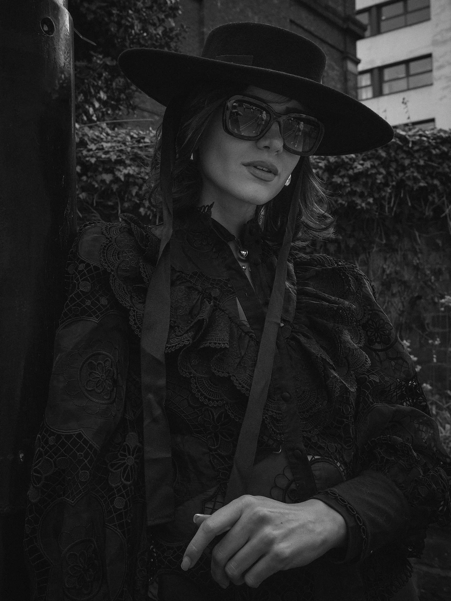 A woman wearing a large wide-brimmed hat, sunglasses, and a lace embroidered jacket, sitting outdoors with her hands clasped, with a background of trees, bushes, and a building.