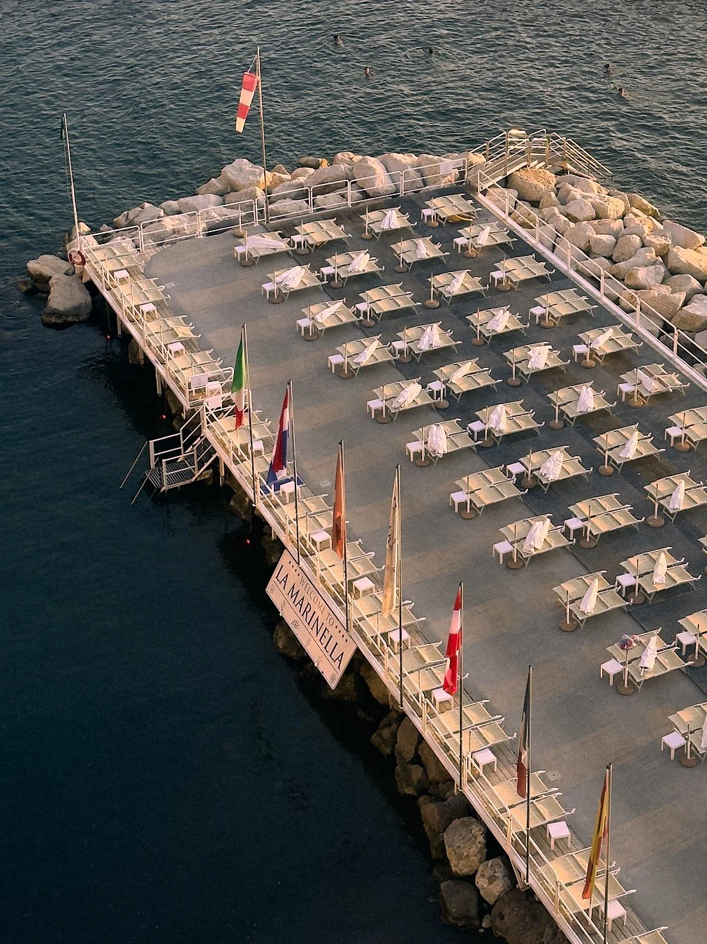 An empty outdoor dining area on a pier called La Marinella, with white tables and chairs covered with white cloths, overlooking the water. Flags are flying along the railing, and there are rocks surrounding the pier.