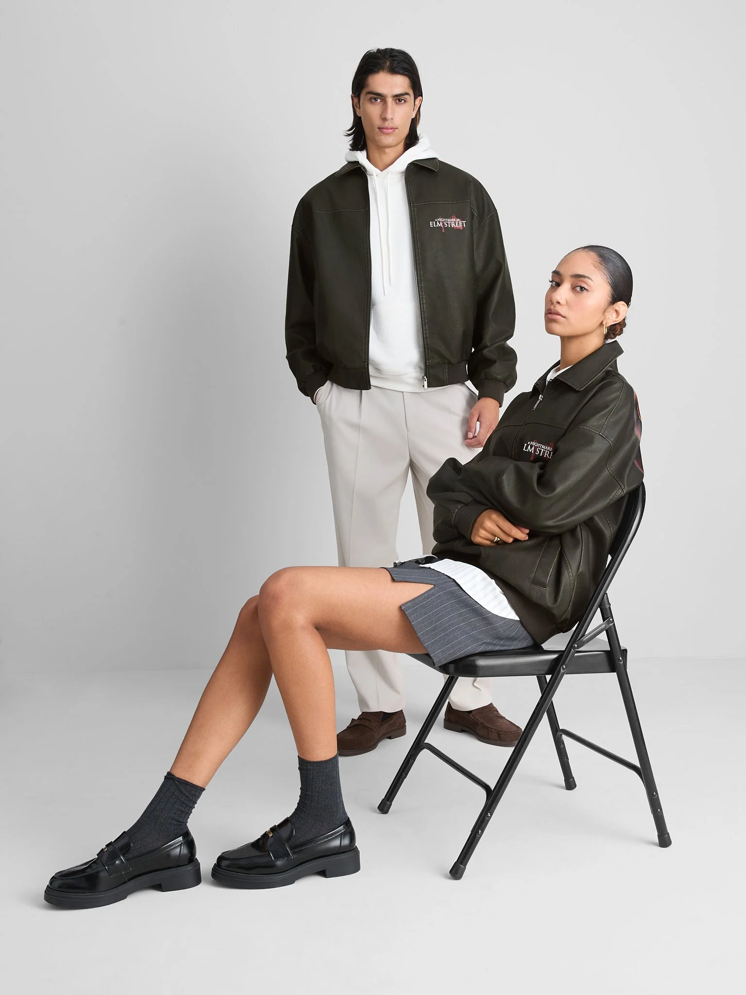Two young fashionably dressed individuals, a man standing and a woman sitting on a black folding chair, pose against a plain gray background. They wear casual clothing with a streetwear style.