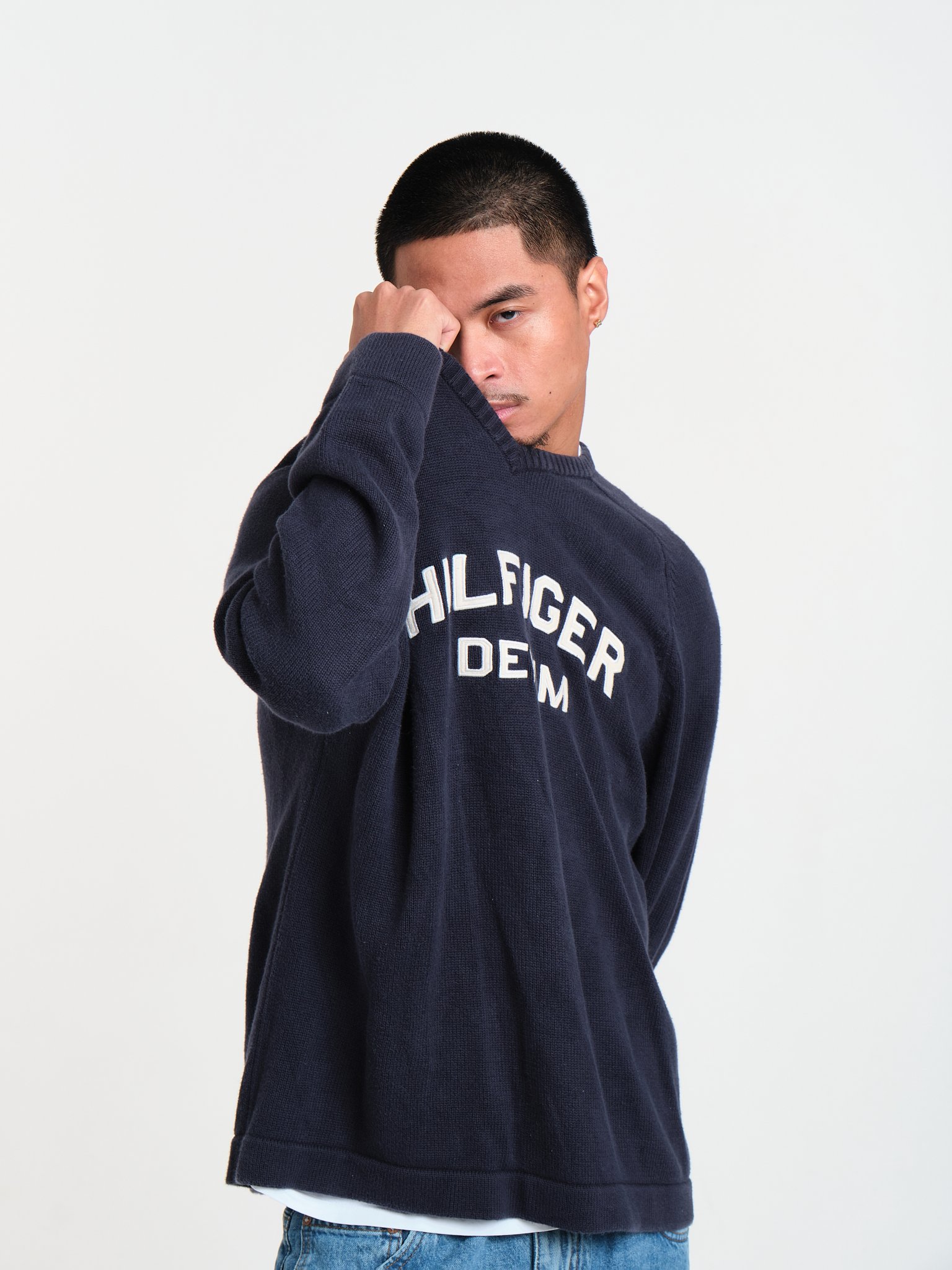 A young man with short black hair, wearing a navy blue Tommy Hilfiger sweatshirt, partially covering his face with his right arm, standing against a plain white background.