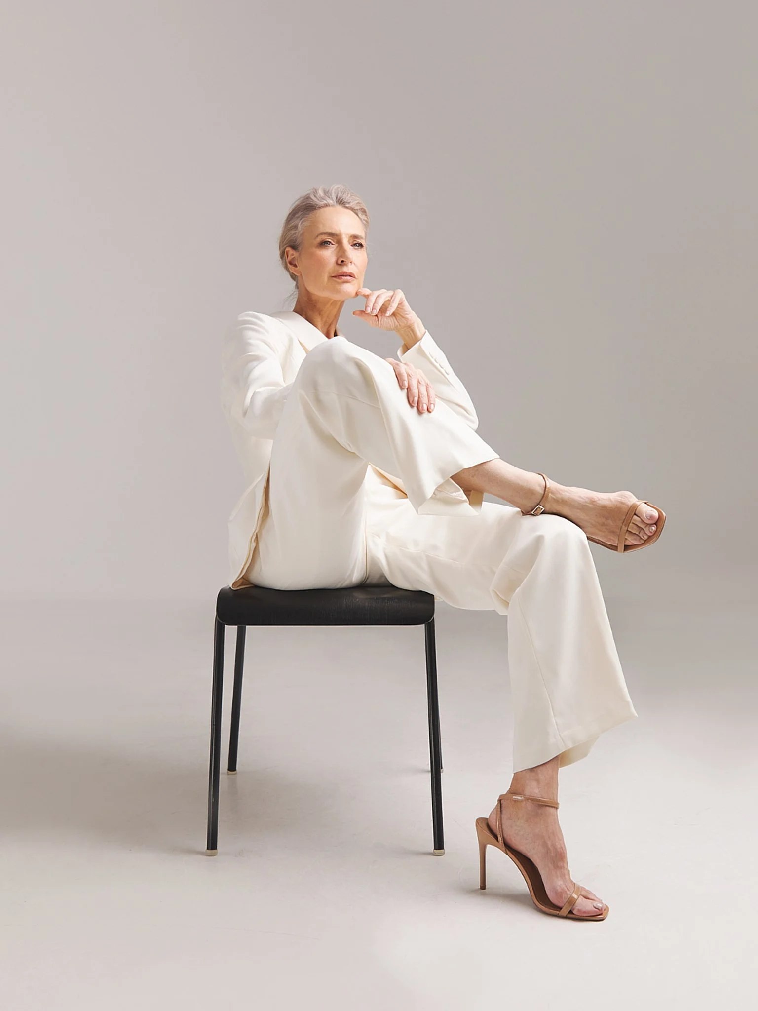 An elderly woman with silver hair styled back, wearing a cream-colored outfit and nude high-heeled sandals, seated on a black chair against a plain light gray background, with a thoughtful expression, sitting with one leg crossed over the other.