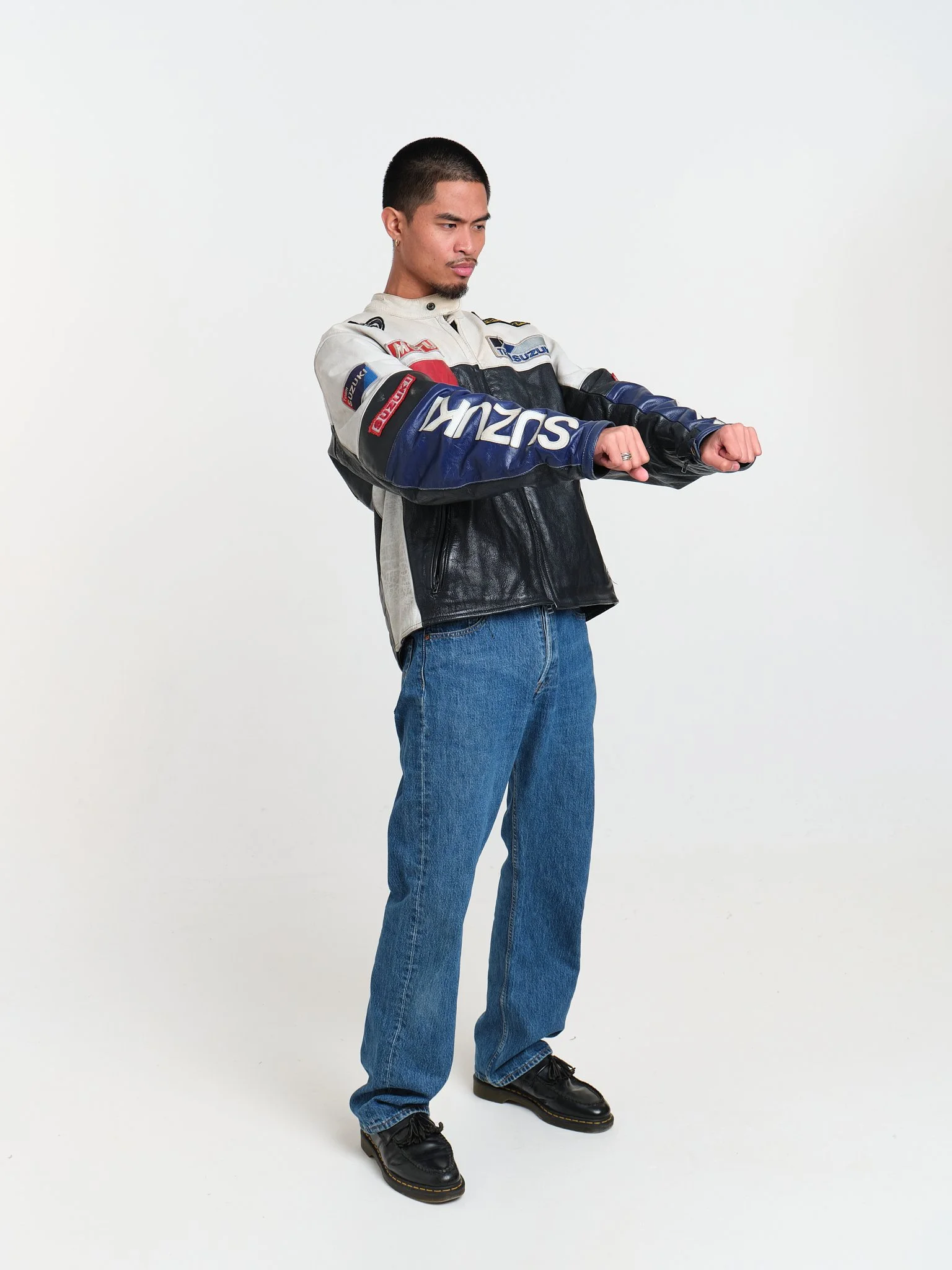 Young man wearing a motorcycle racing jacket and jeans standing against a plain white background, looking at his watch.