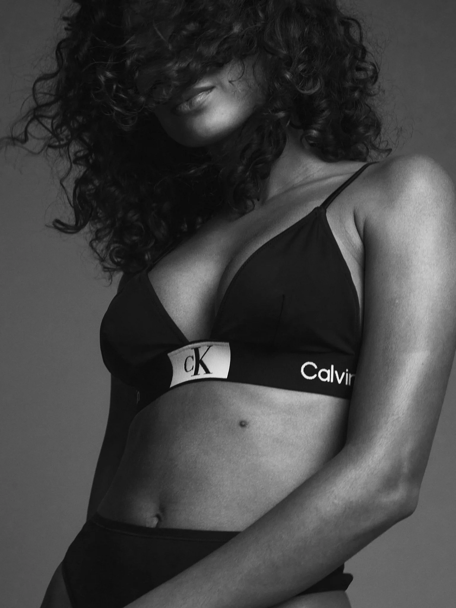 Close-up black-and-white photo of a woman with curly hair wearing a Calvin Klein bra, showing part of her face and torso.