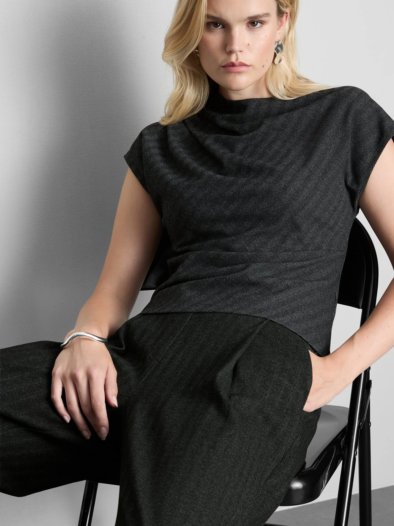 A woman sitting in a black chair, wearing a black textured short-sleeve top and black pants, with her right hand resting on her lap and her left hand on her hip. She has blonde hair, light skin, and is wearing silver earrings and a silver bracelet.