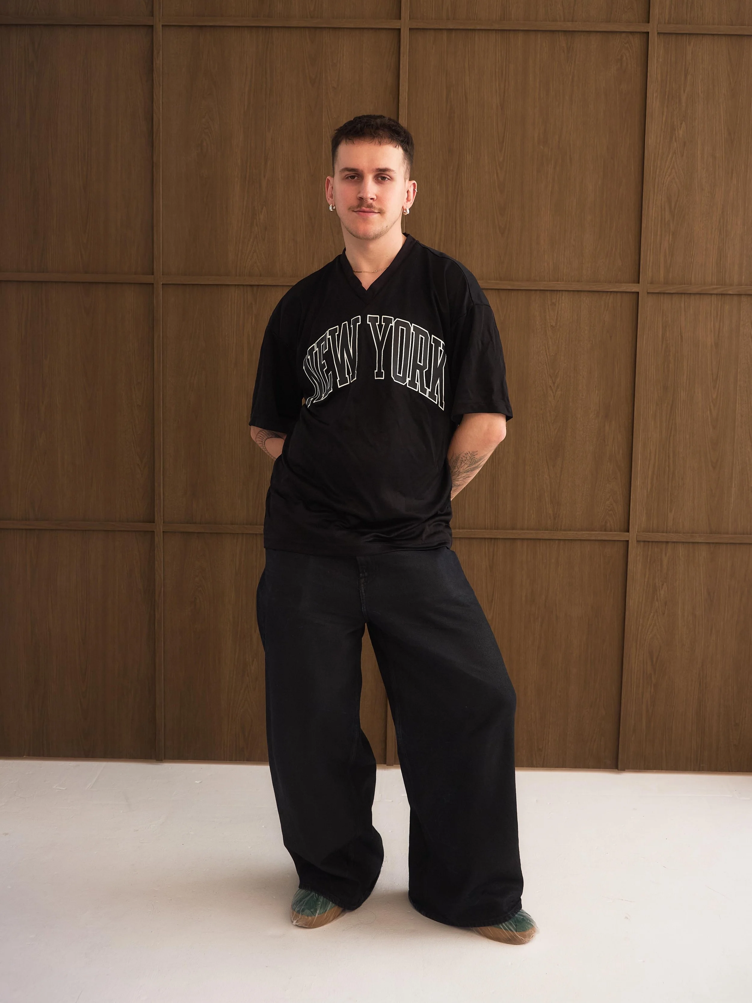 A young man with short brown hair, earrings, and tattoos on his forearms, wearing a black T-shirt with 'New York' written on it, dark pants, and green slippers, standing against a wooden wall.