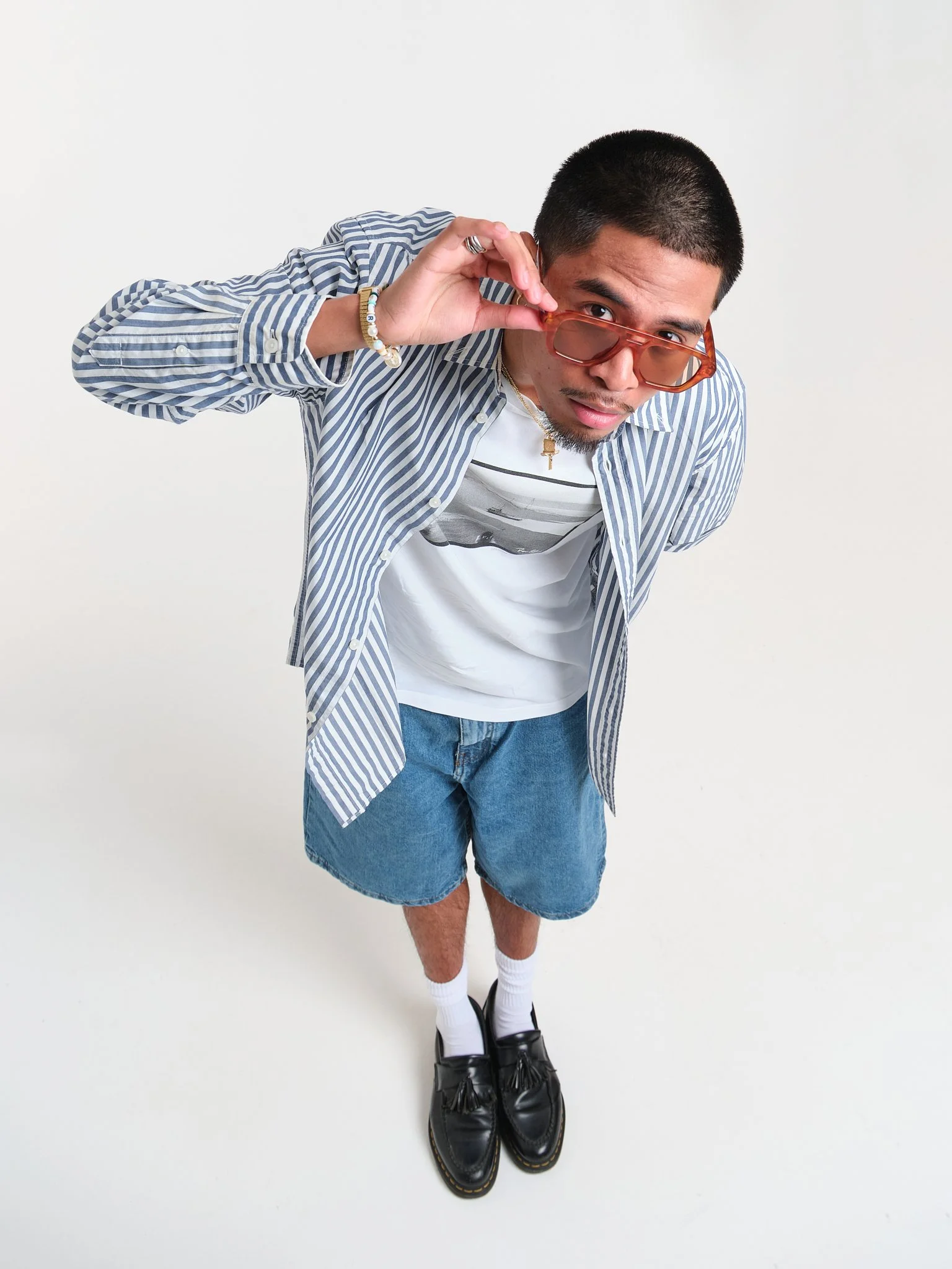 Young man with short hair, wearing sunglasses, striped shirt, white t-shirt, denim shorts, white socks, black shoes, holding sunglasses, standing against a plain white background.