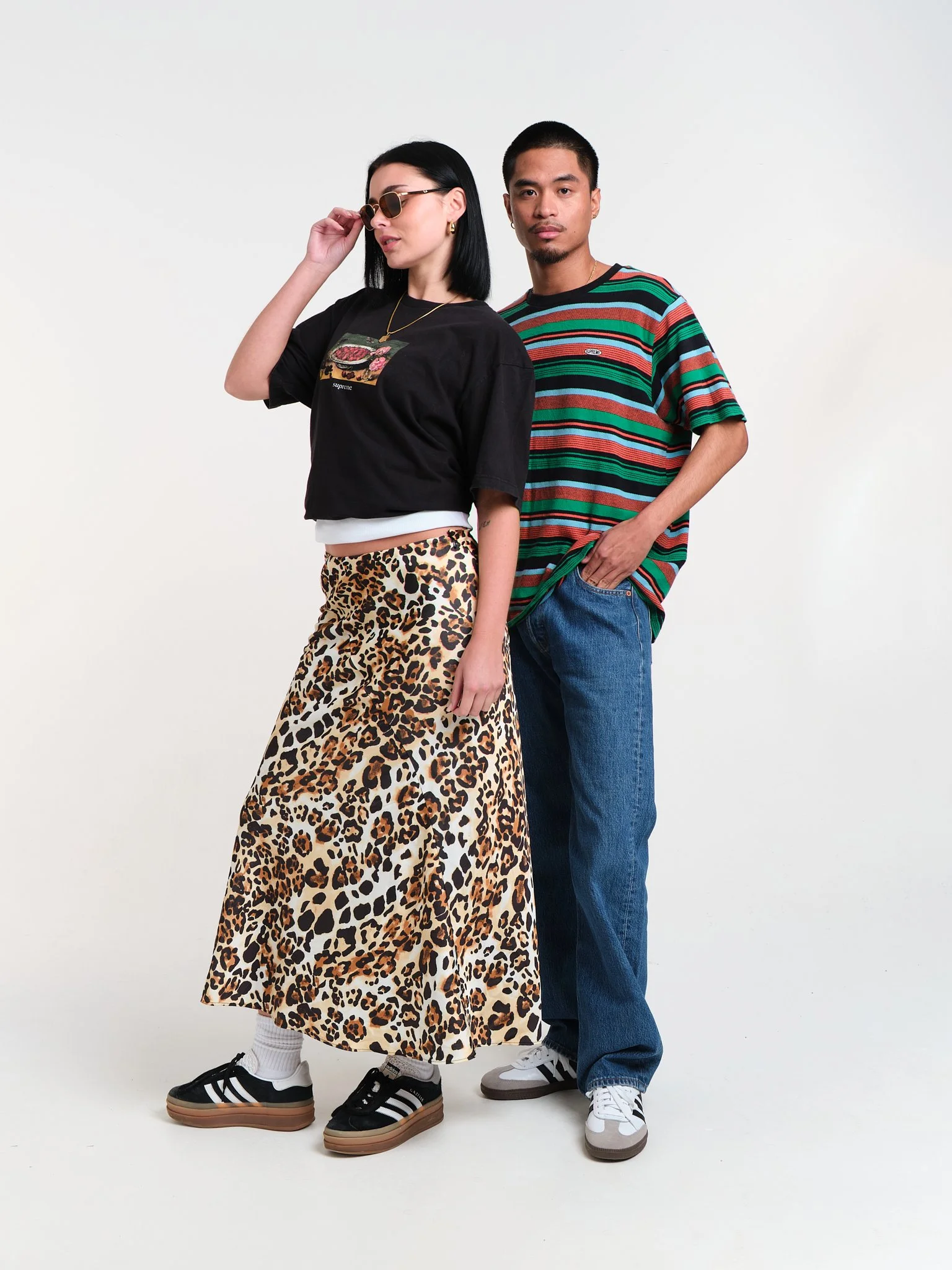 Two young adults, a woman and a man, stand against a plain white background. The woman is wearing sunglasses, a black T-shirt with a graphic, a white undershirt, a leopard print skirt, and black sneakers. The man is dressed in a multicolored striped 