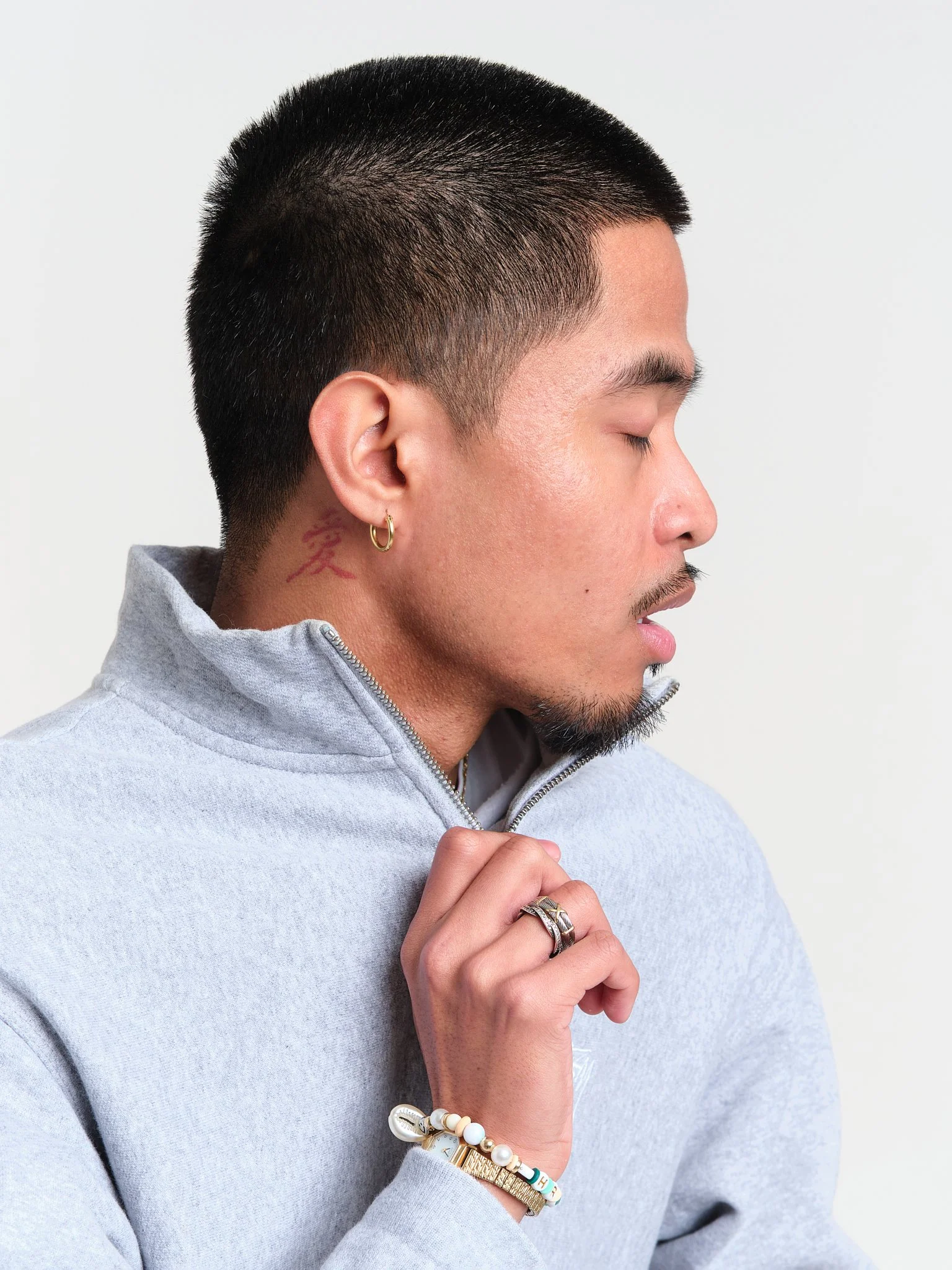 Profile of a young woman with short black hair, gold hoop earrings, and tattoos on her neck, wearing a gray zip-up sweater, with her eyes closed and her hand gently resting on her chest, against a plain background.