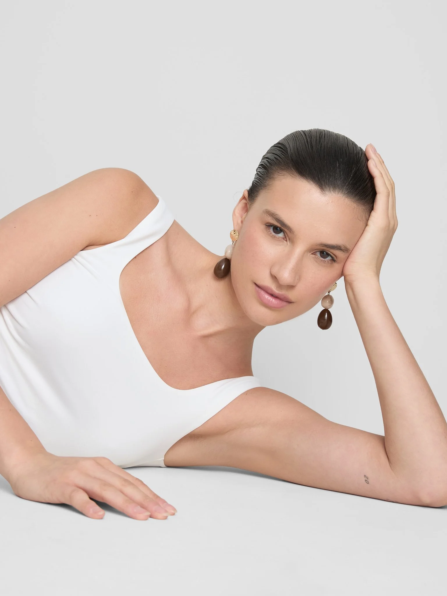 A woman lying on her side with her head resting on her hand, wearing a white sleeveless top and colorful earrings, against a plain light gray background.