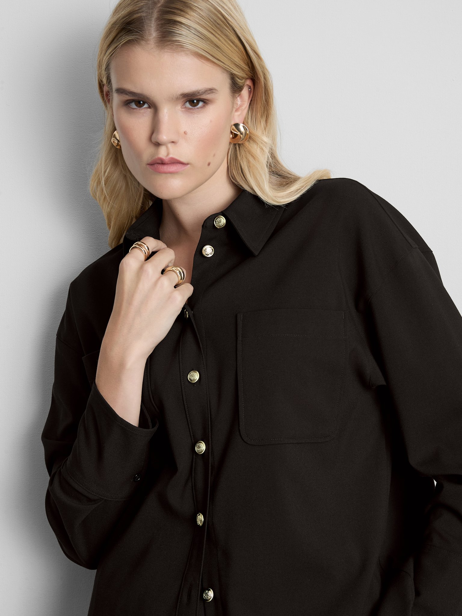 A woman with blonde hair wearing a black button-up shirt with gold buttons, gold earrings, and gold rings, standing against a plain gray background.