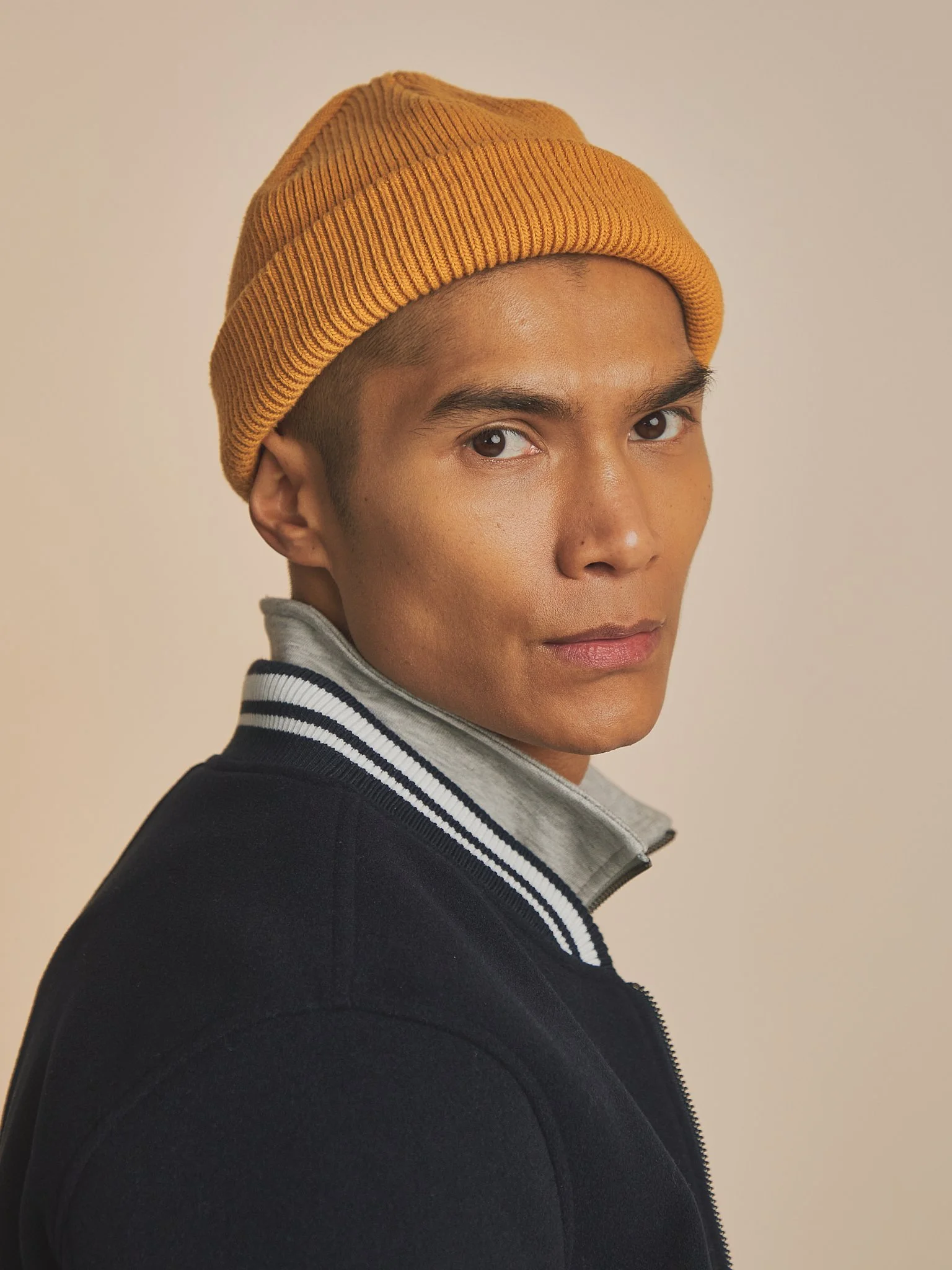 Close-up of a young person with short hair wearing a mustard yellow knit beanie and a black fleece jacket with a gray and white striped collar, against a plain beige background.