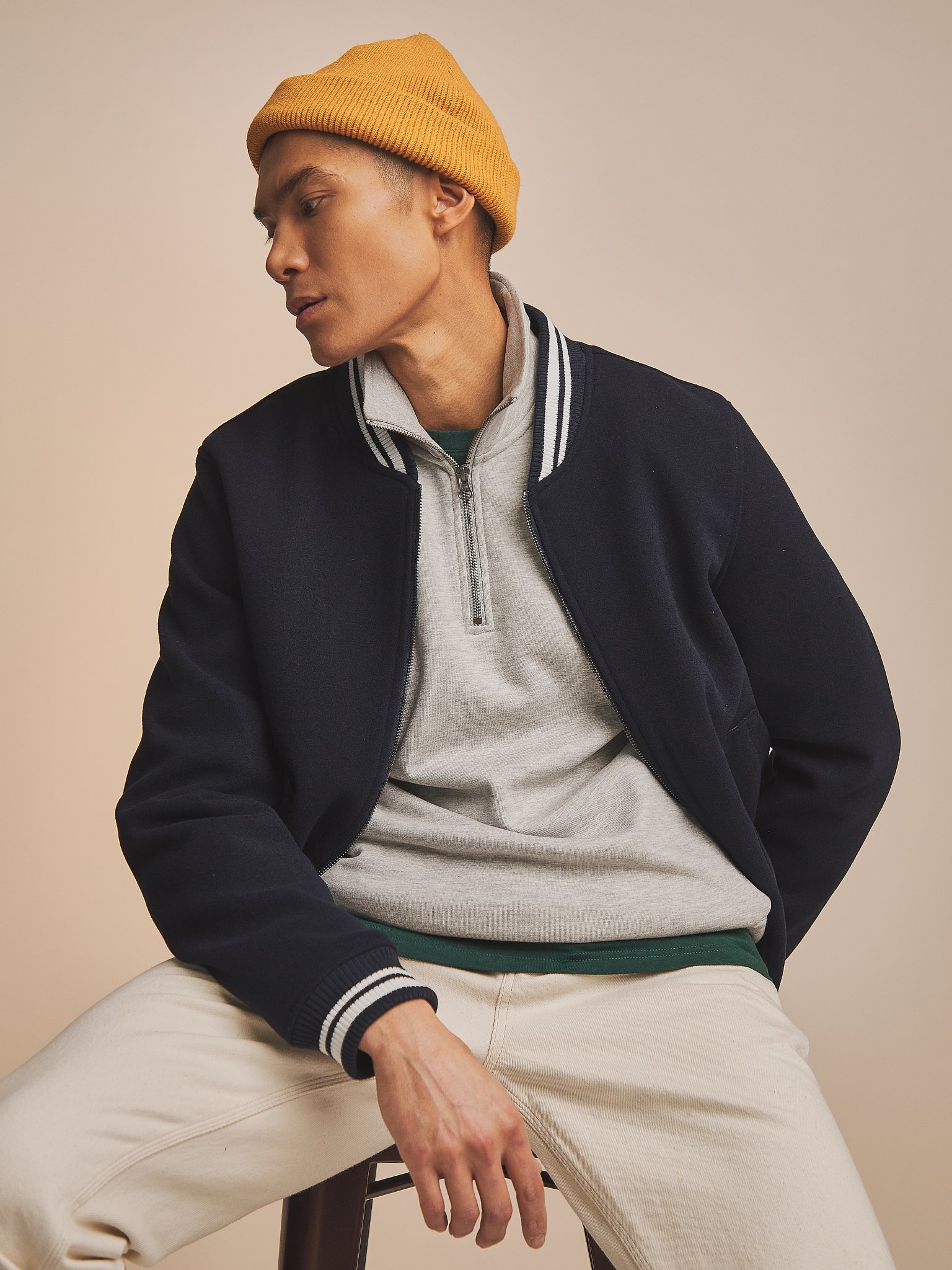 A young man wearing a mustard yellow beanie, a navy blue varsity jacket, a gray zip-up sweater, and beige pants, sitting on a stool against a plain beige background.