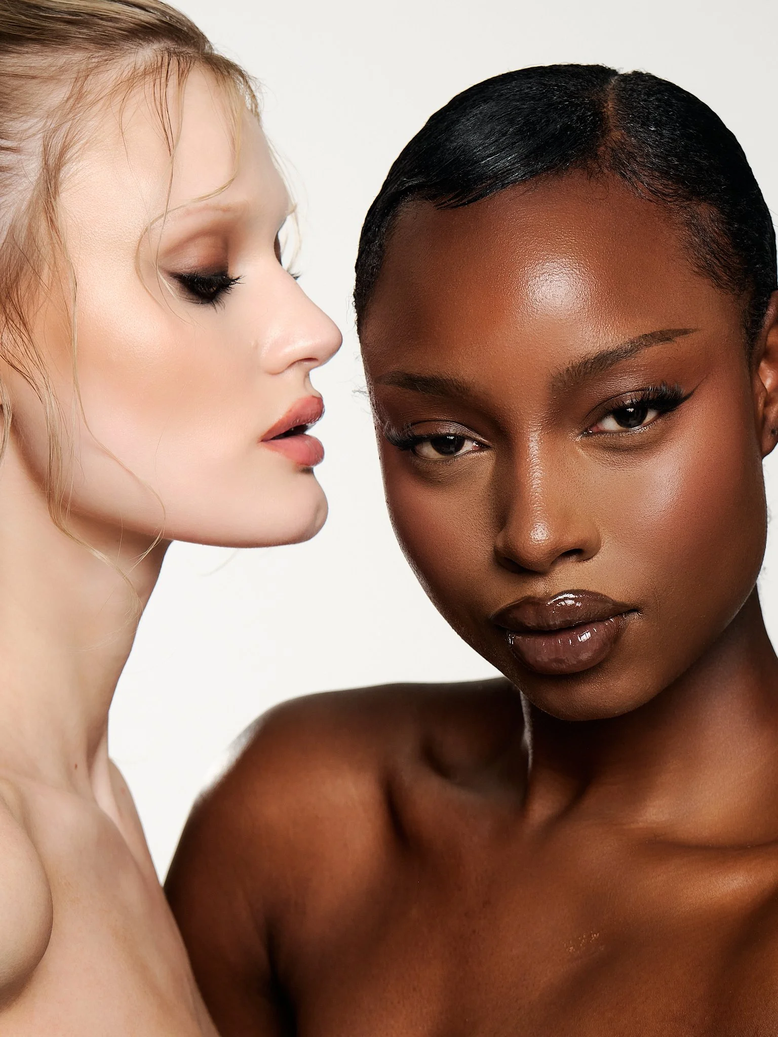 Close-up of two women with different skin tones and hair colors, facing each other against a white background.