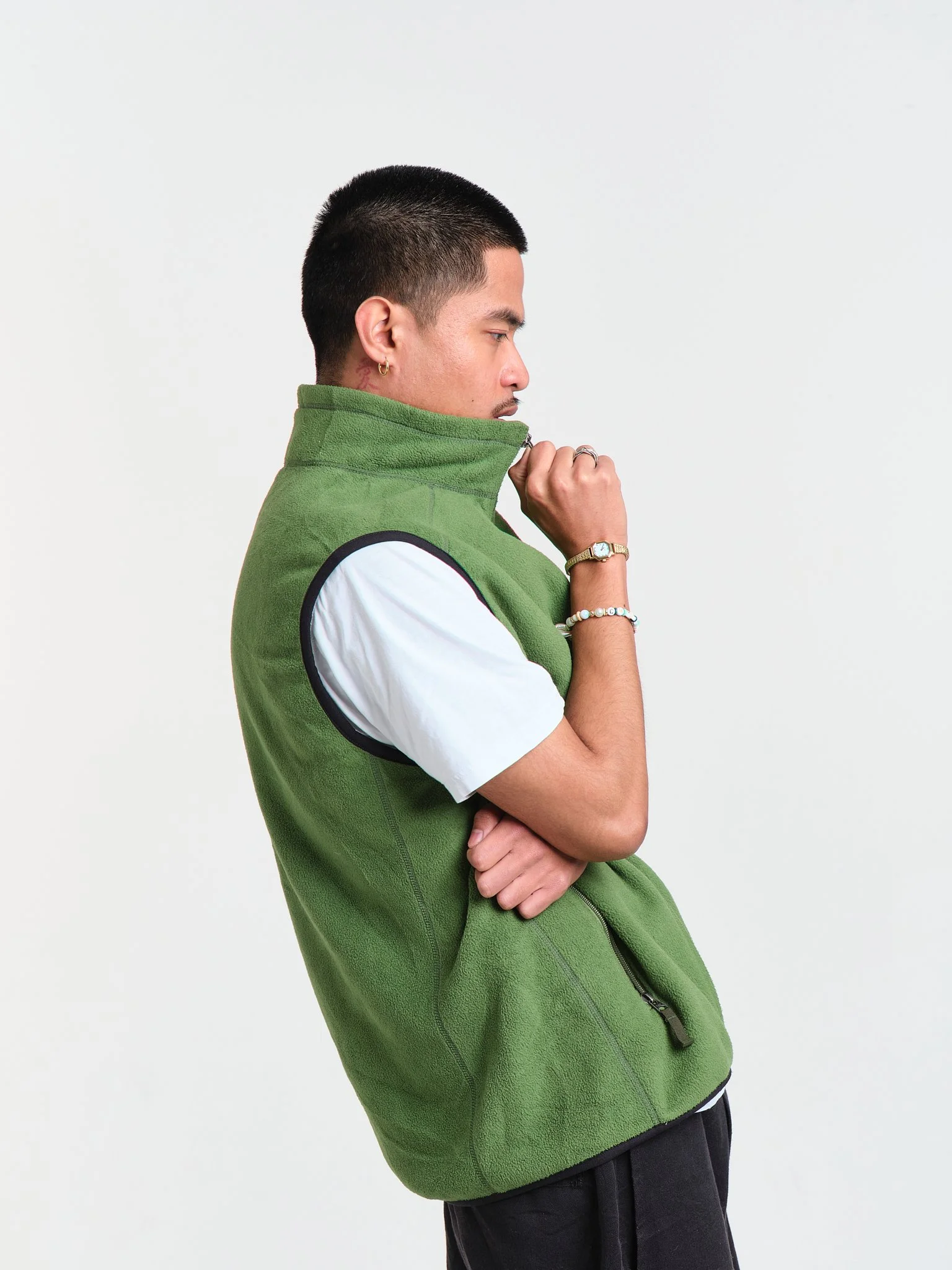 A young man with short black hair and earrings, wearing a green fleece vest over a white T-shirt, with his eyes closed, standing against a plain white background, holding the collar of his vest with one hand and crossing his arms.