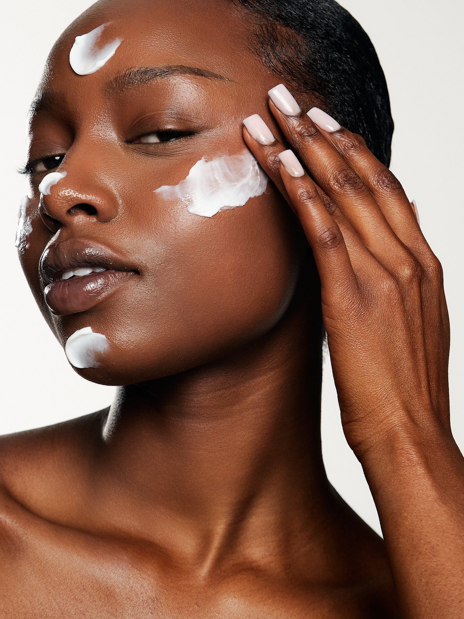 A close-up of a woman with dark skin applying white cream to her face, with her hand near her cheek and eye area, against a plain light background.