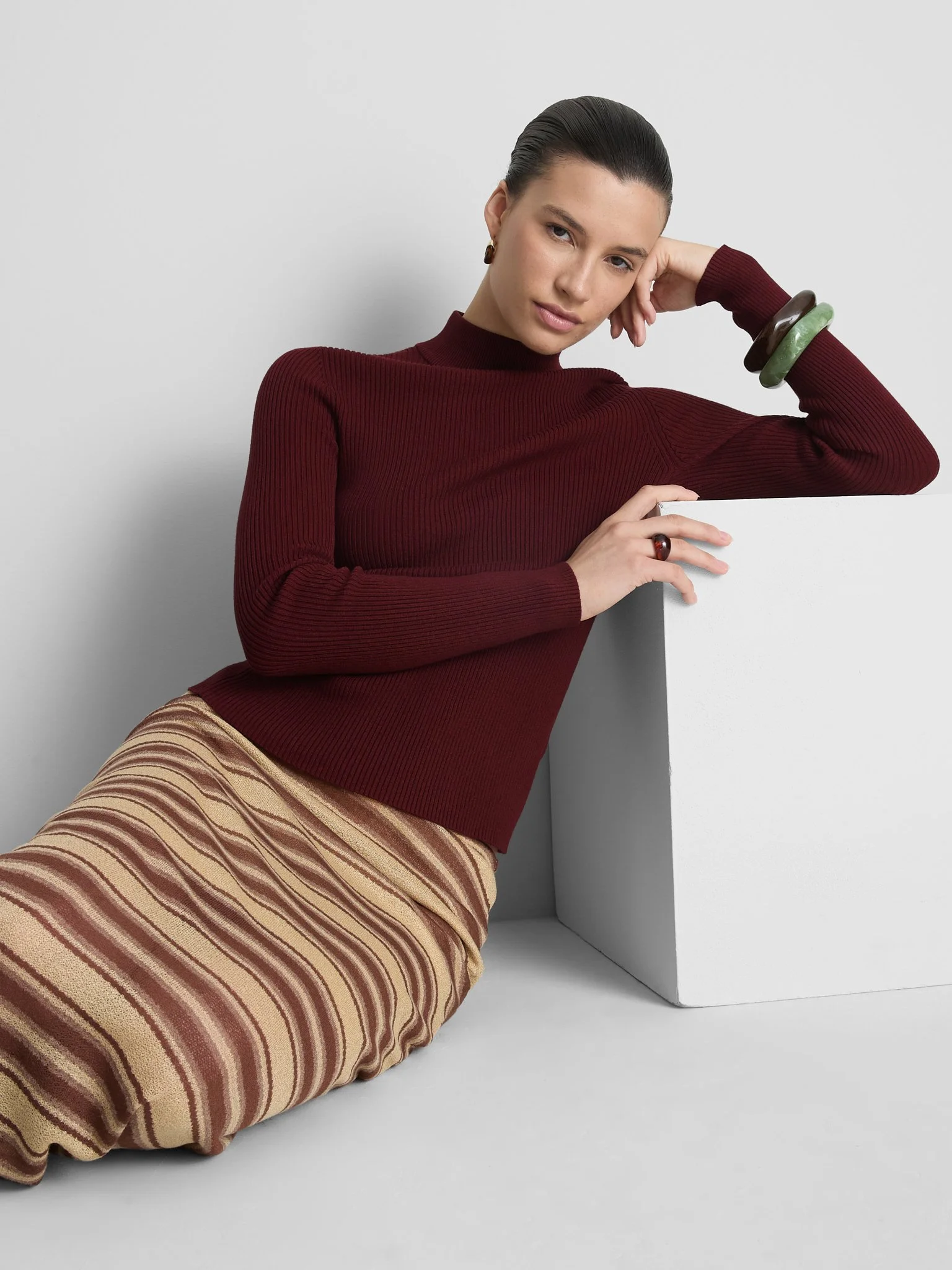A woman with dark hair slicked back, wearing a red long-sleeve sweater, striped skirt, and colorful bracelets, leaning against a white surface, looking at the camera.