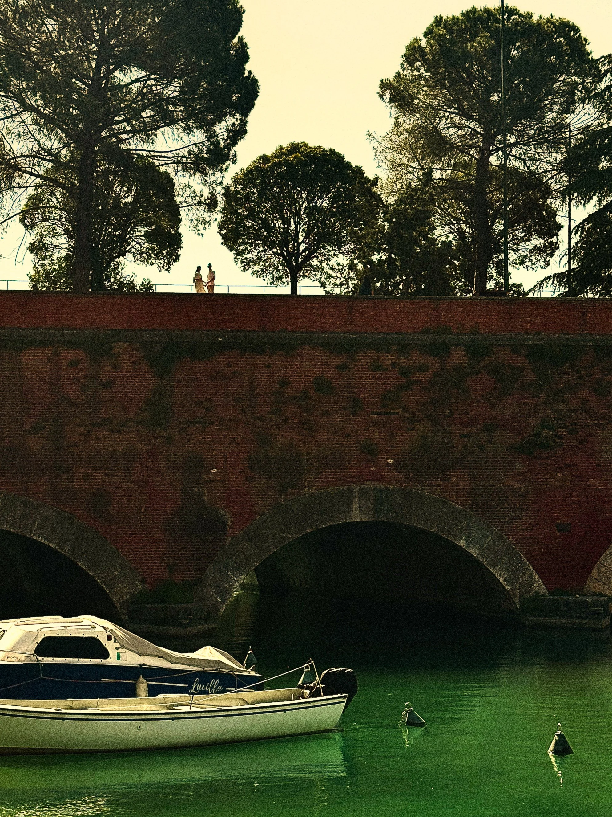 A boat named 'Lumilla' docked on a river beneath a brick bridge with arches, trees on the bank, and boats moored nearby.