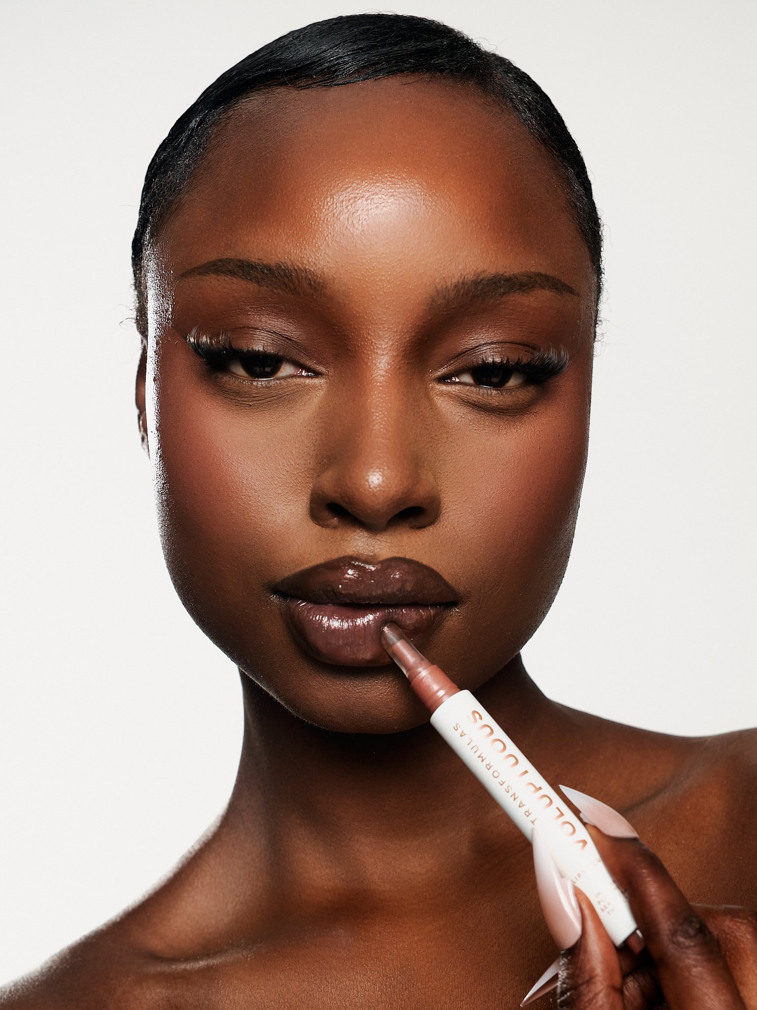 A close-up of a woman with dark skin and sleek hairstyle applying a nude lipstick with a white and pink applicator.