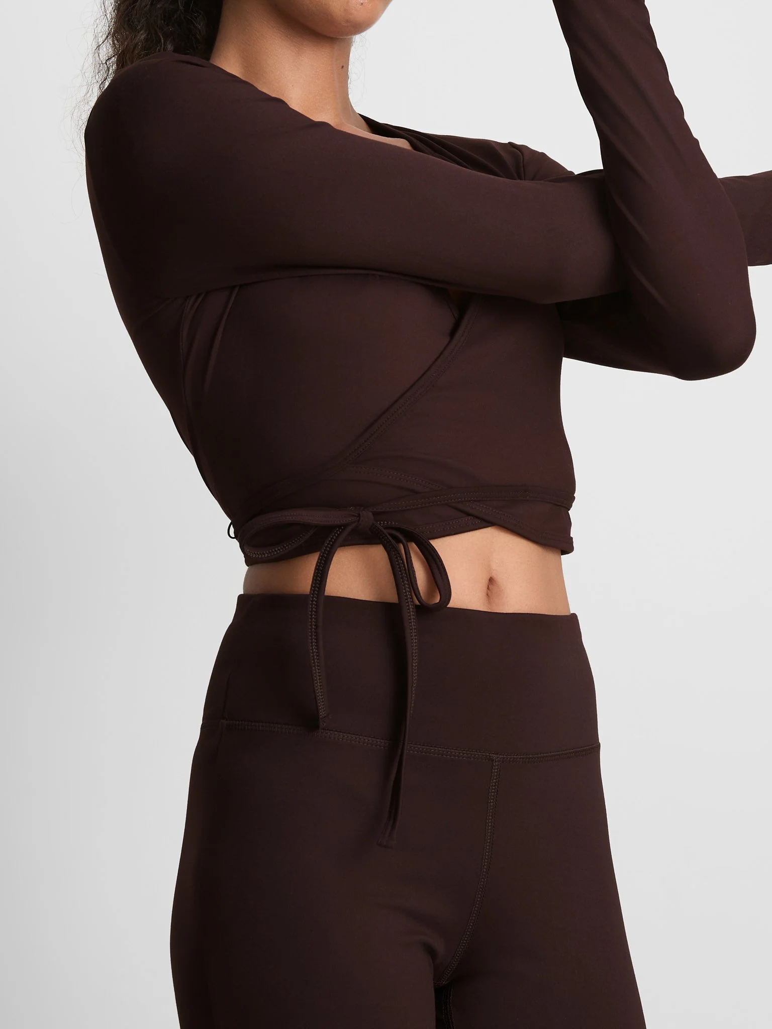 Close-up of a woman wearing a dark brown long-sleeve top and matching dark brown athletic pants, with her arms crossed and hands resting on her shoulders against a plain light background.