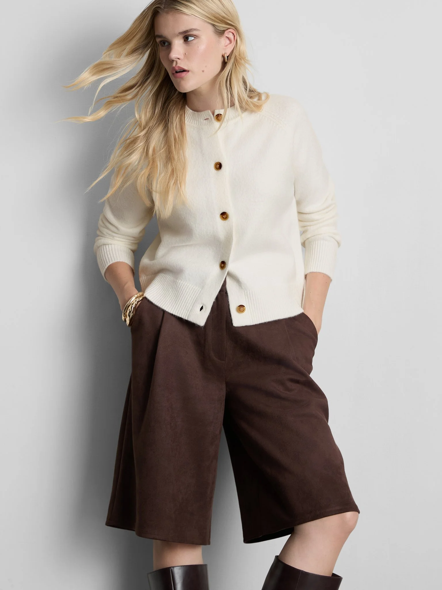 A young woman with blonde hair wearing a cream-colored cardigan with brown buttons and brown wide-leg pants, standing against a gray wall.