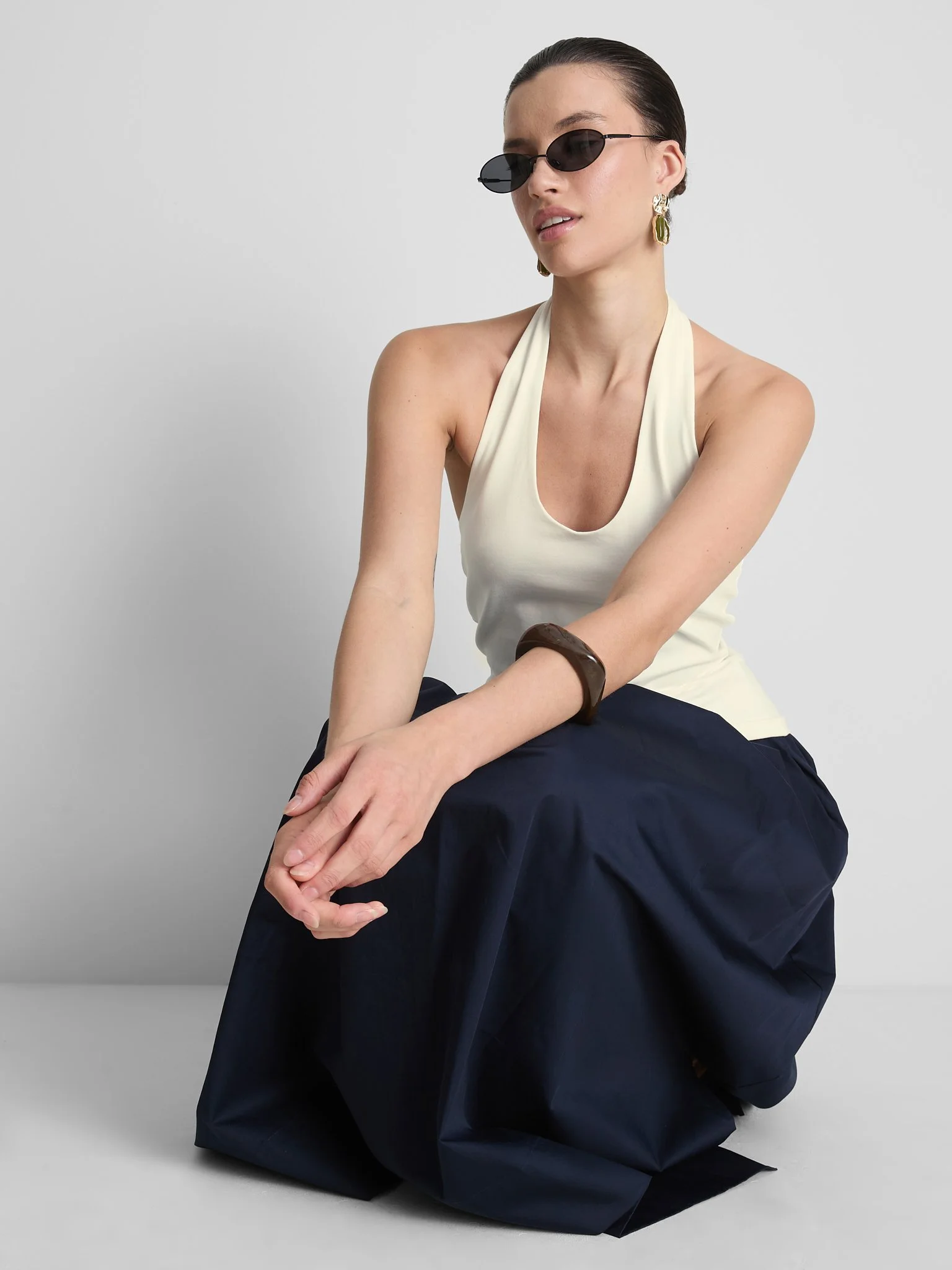 A woman sitting on the floor with her knees bent, wearing sunglasses, a cream-colored sleeveless top, large earrings, a dark bracelet, and navy wide-leg pants against a plain light gray background.