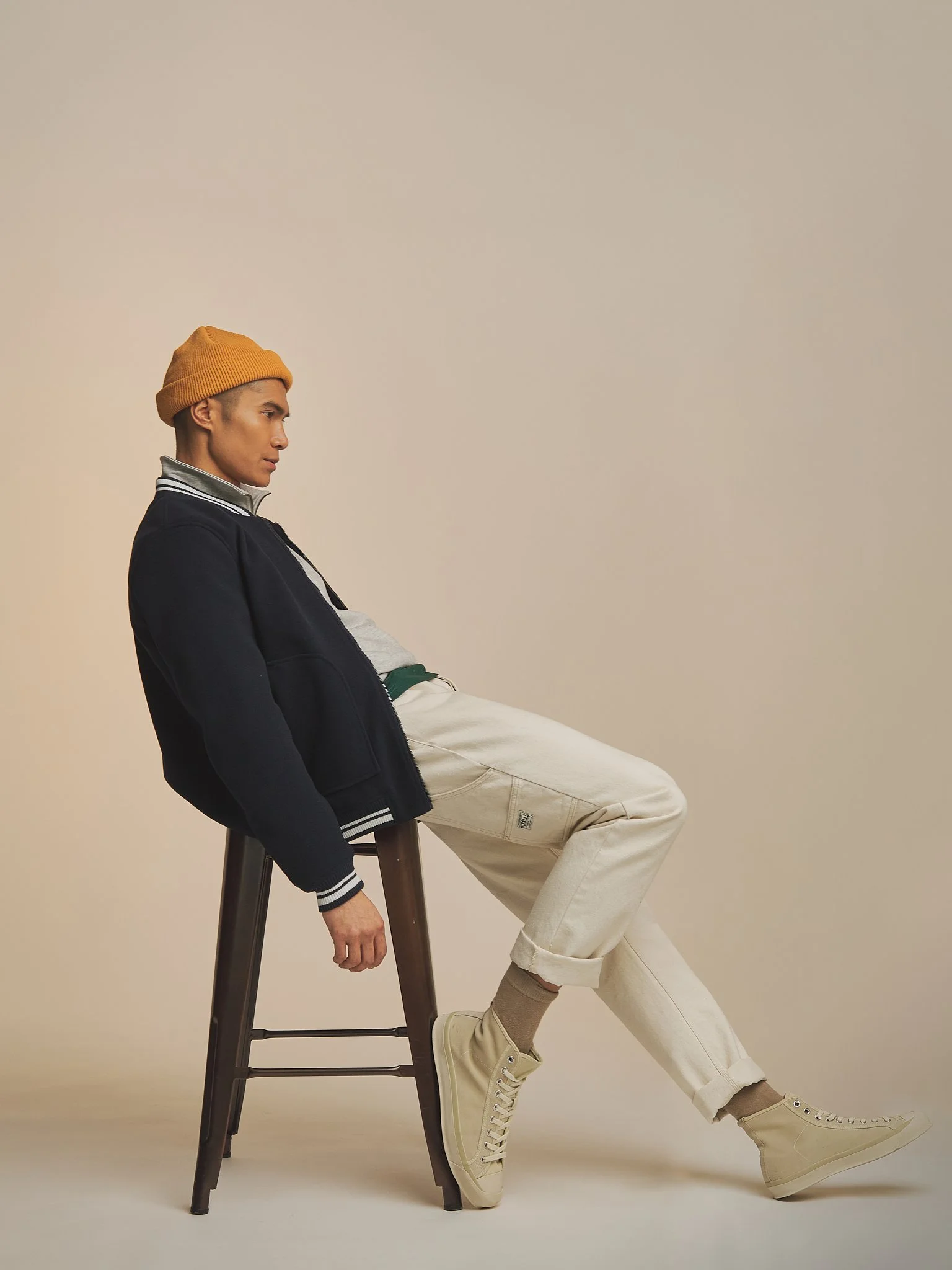 A young man with a yellow beanie and a navy and white varsity jacket sitting on a stool, leaning on a wooden ladder with one leg up, wearing beige pants and sneakers, against a plain light background.