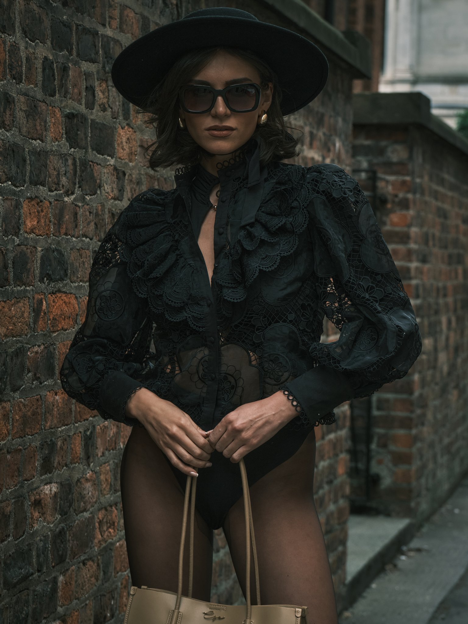 A woman wearing a black hat, large sunglasses, and a black lace shirt with puffed sleeves, standing against a brick wall in an urban setting.